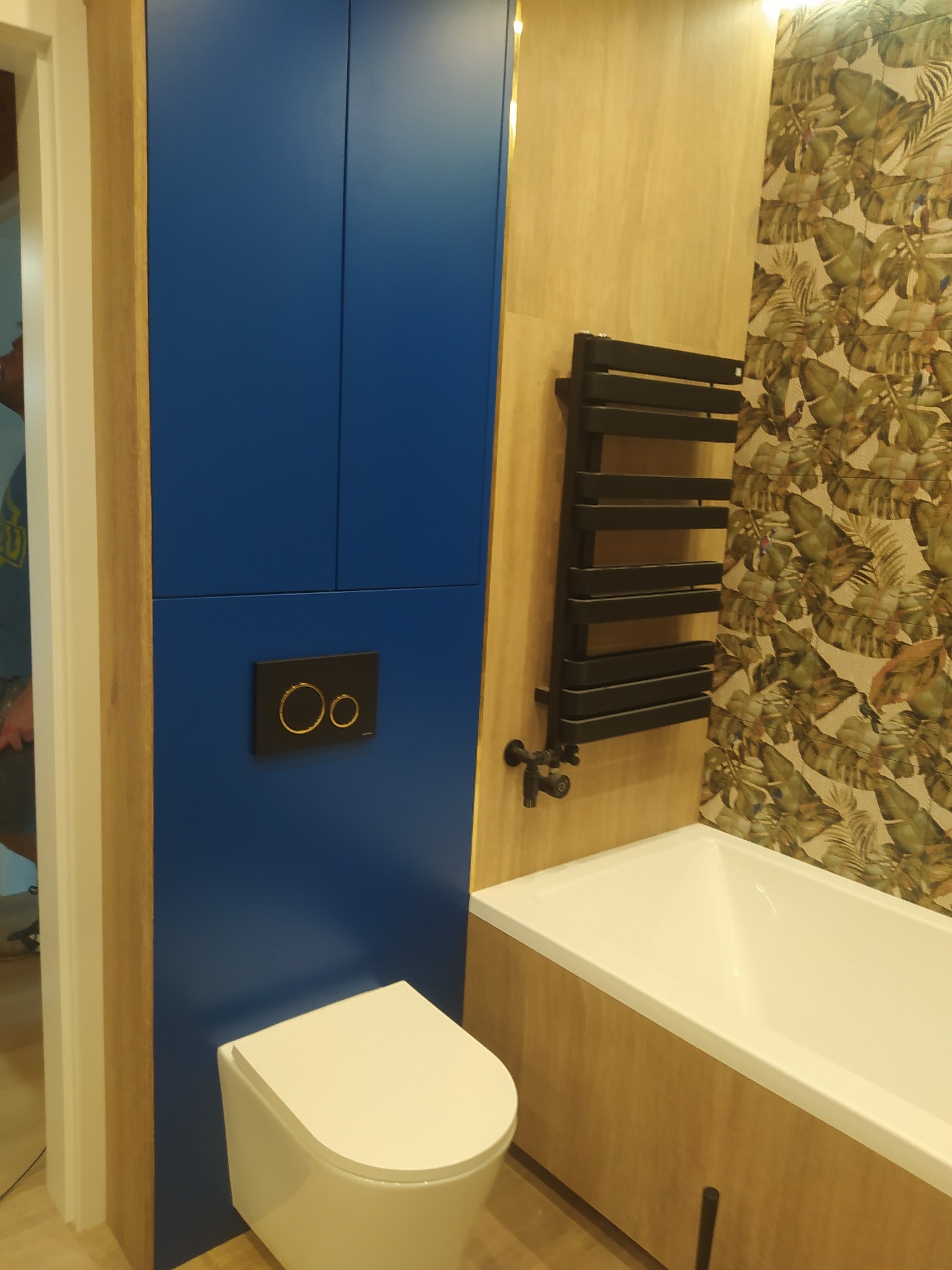 Nowoczesna łazienka z zabudowaną toaletą w niebieskiej szafce, grzejnikiem drabinkowym i wanną obudowaną drewnopodobną płytą z motywem liści.