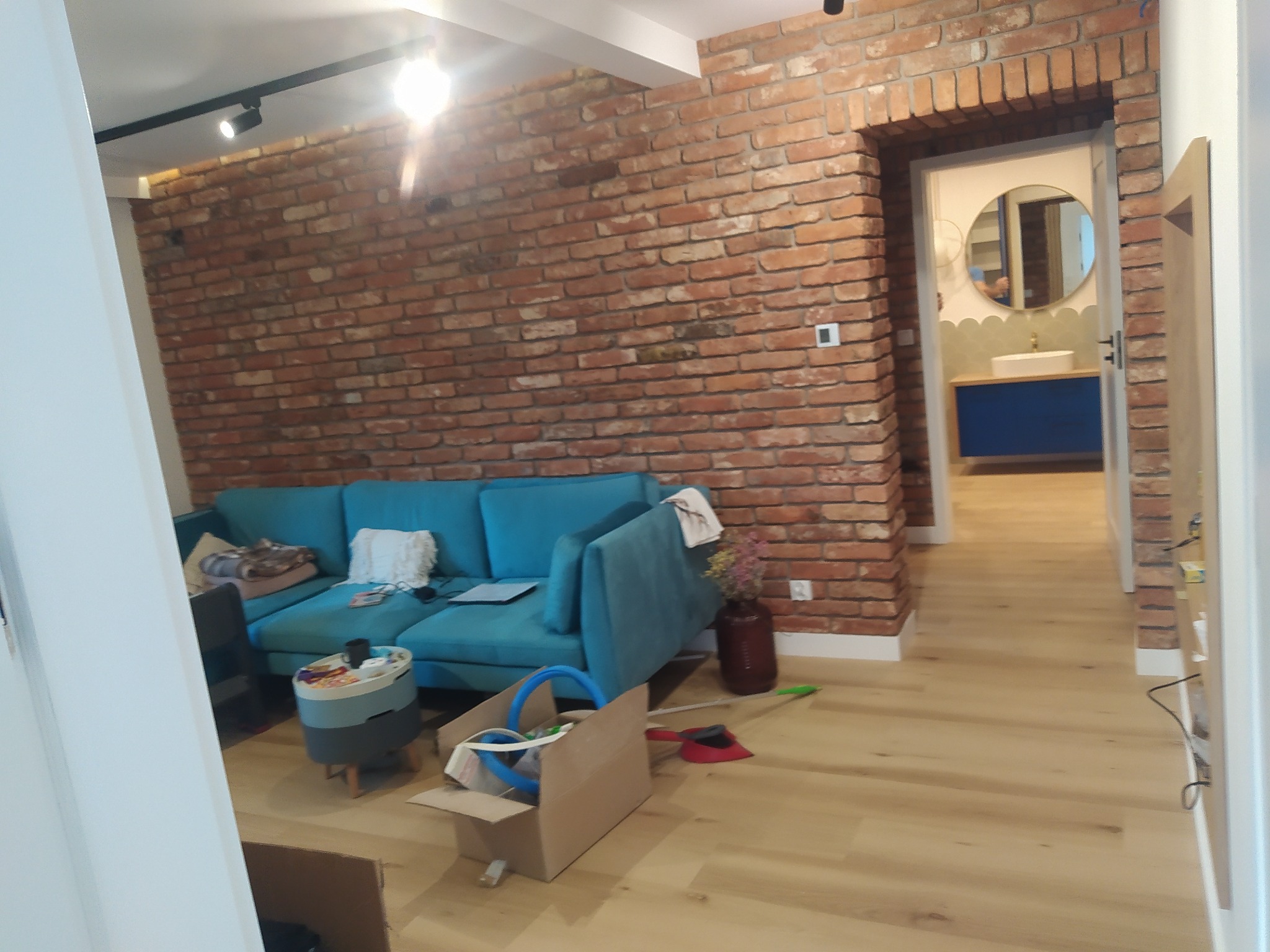 Salon z ceglaną ścianą i turkusową sofą, widok na łazienkę z okrągłym lustrem, częściowo posprzątane po remoncie, karton z narzędziami na podłodze.