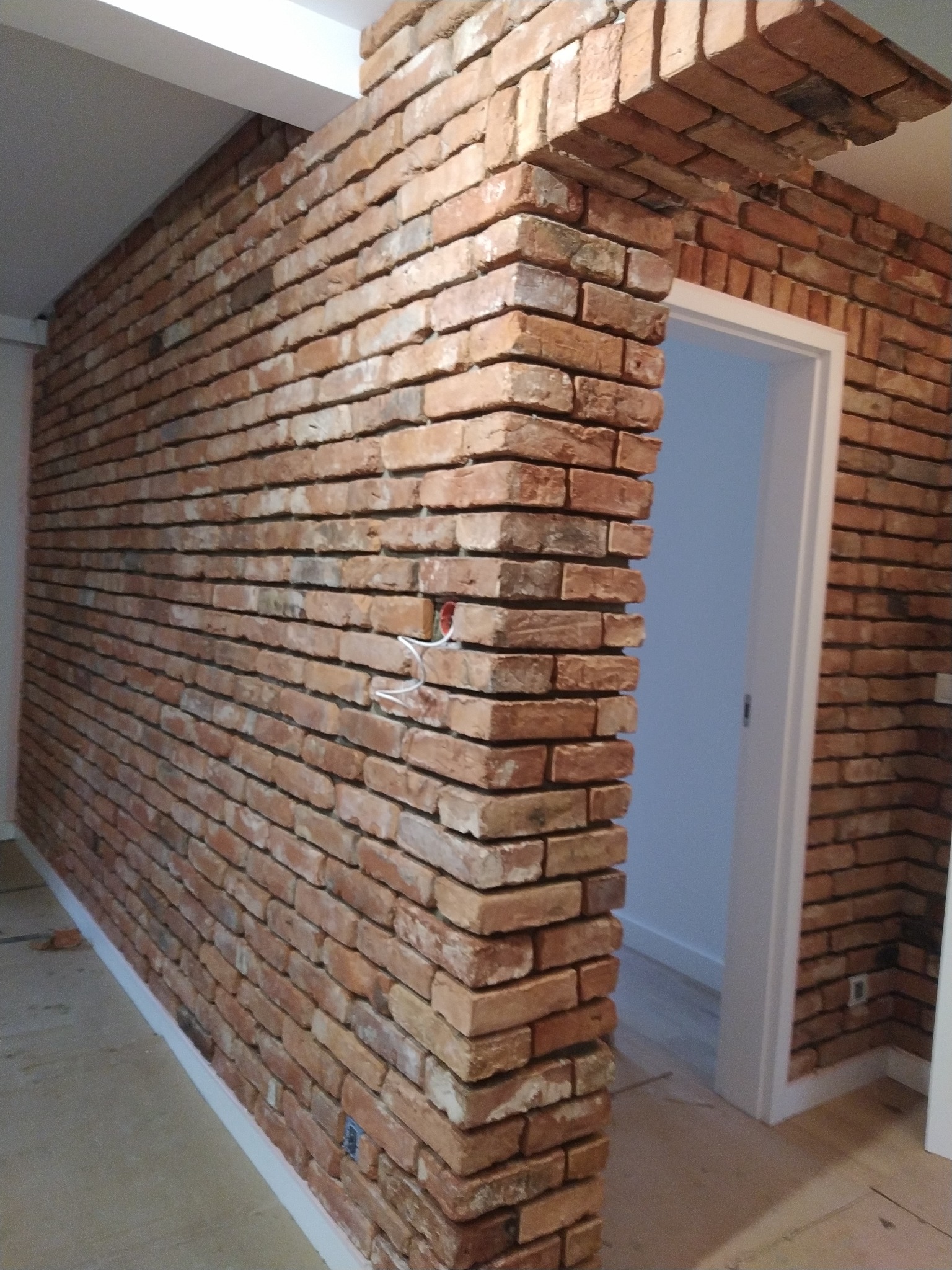 Ściana z czerwonej cegły klinkierowej wewnątrz pomieszczenia, widoczne gniazdko elektryczne i fragment podłogi w trakcie prac wykończeniowych.