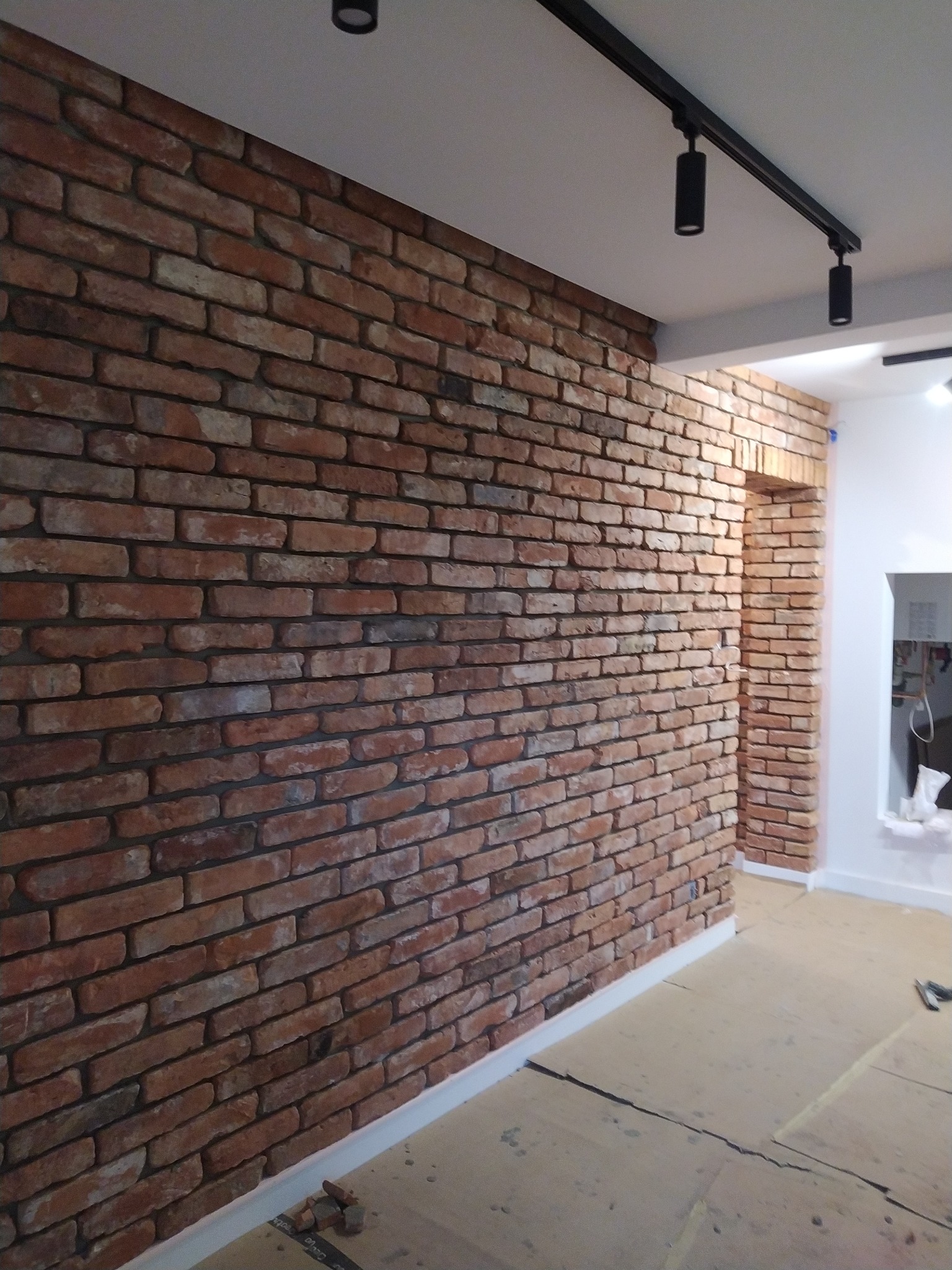 Ściana wykończona czerwoną cegłą klinkierową z widocznym fragmentem podłogi w trakcie prac wykończeniowych i oświetleniem szynowym.