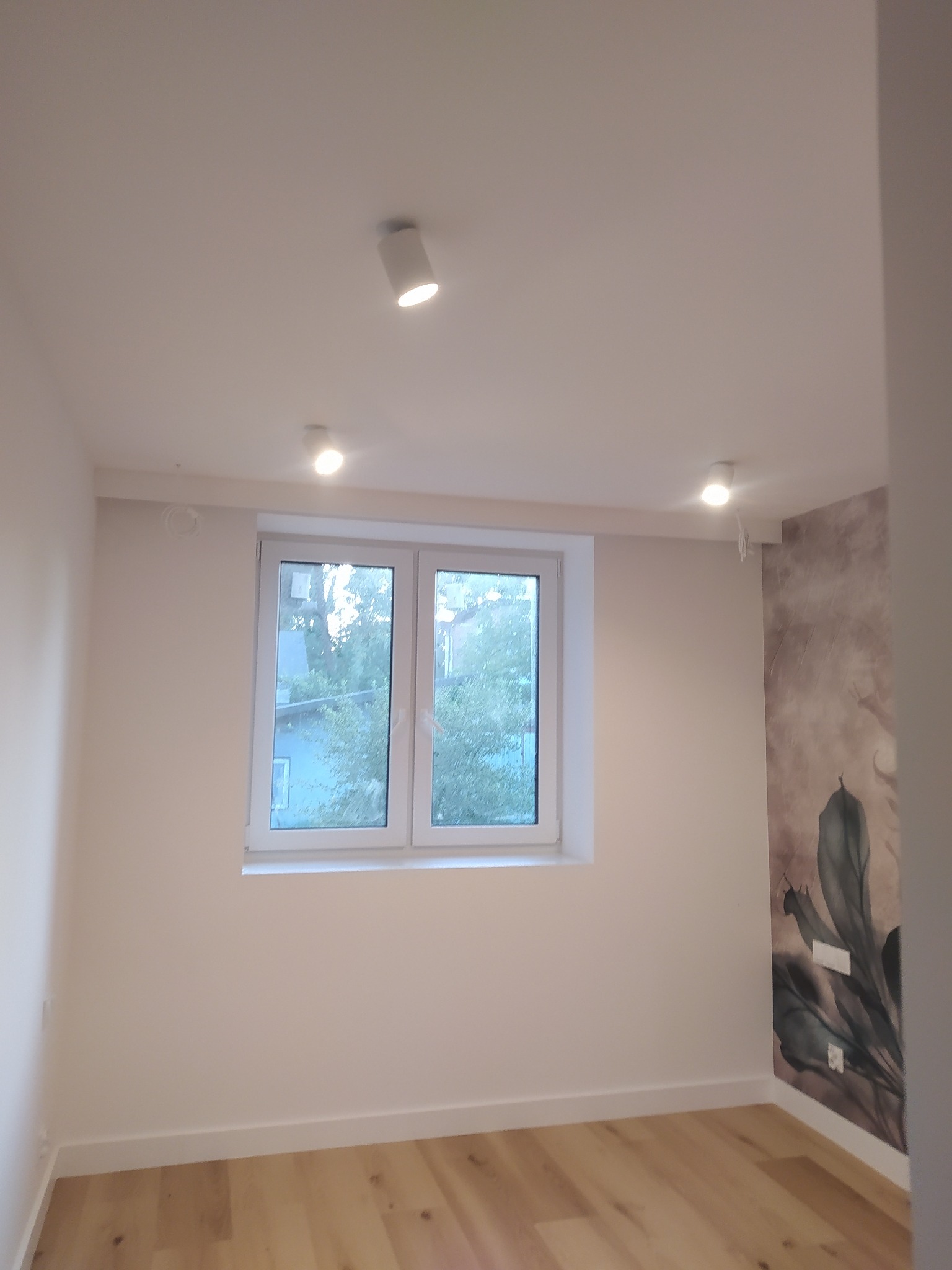 Jasne wnętrze po remoncie z drewnianą podłogą, świeżo pomalowanymi ścianami i nowoczesnym oświetleniem punktowym. Na jednej ze ścian widoczna tapeta z motywem roślinnym.