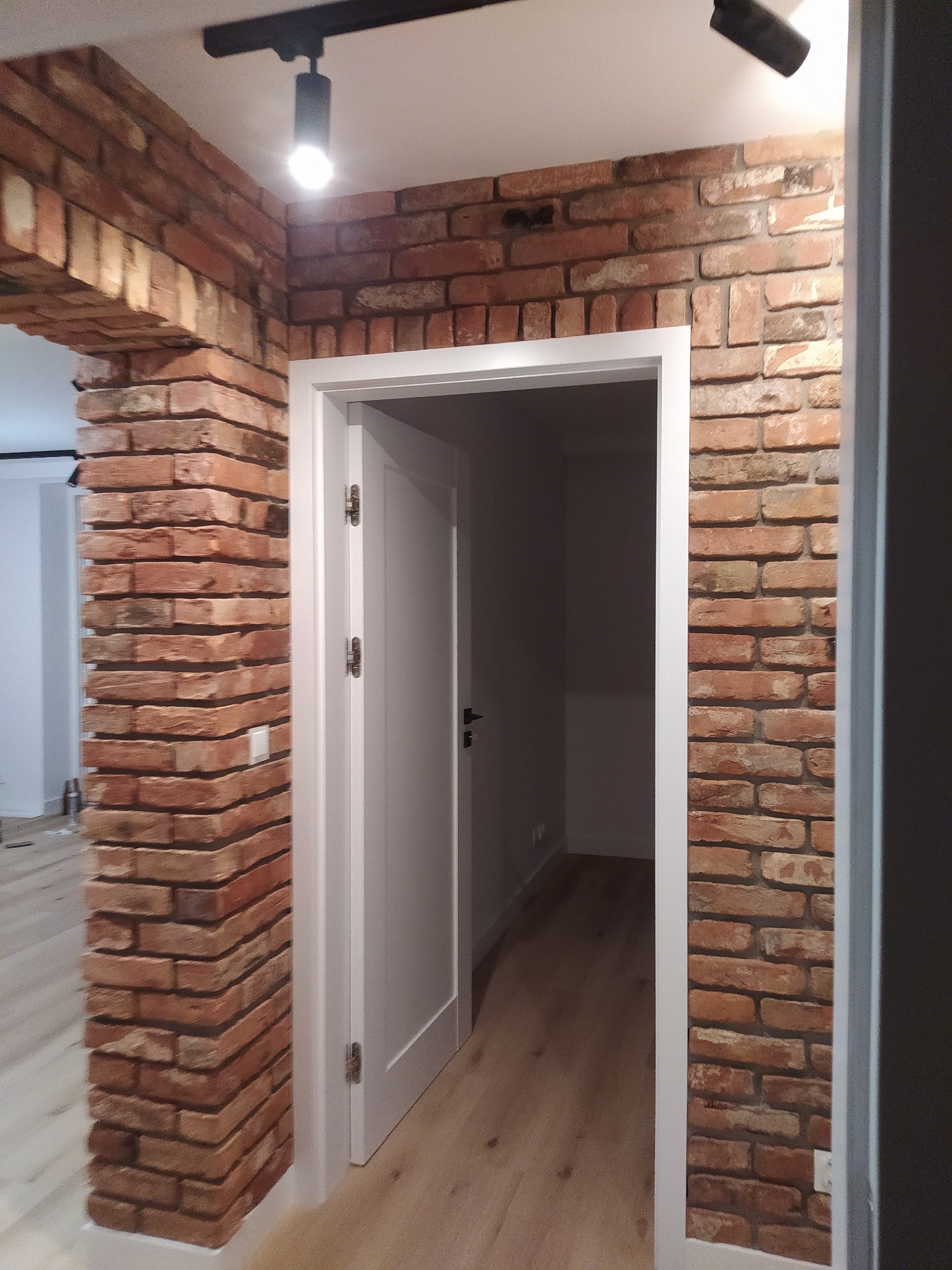 Wnętrze z dekoracyjną ścianą z cegły klinkierowej, oświetleniem punktowym i białymi drzwiami w białej futrynie, widok na parkiet.