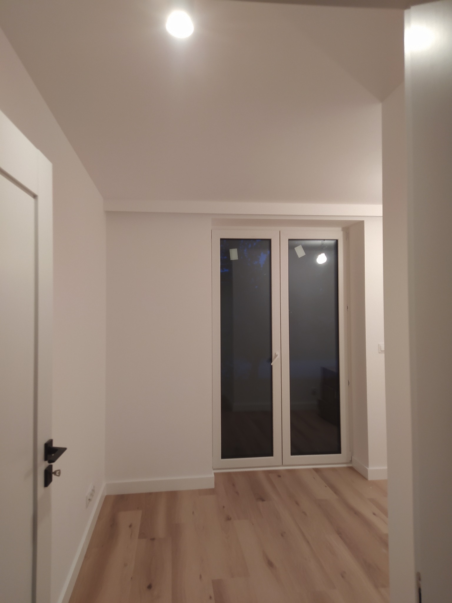 Jasne, nowocześnie wykończone wnętrze pokoju z panelami podłogowymi i białymi ścianami, widoczne białe drzwi z czarną klamką oraz okno balkonowe z naklejkami ochronnymi.