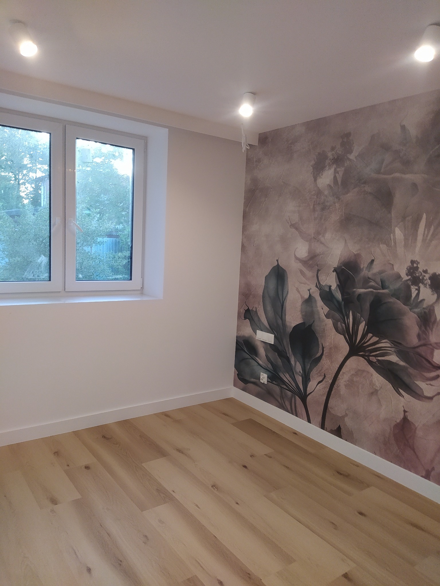 Jasny pokój po odświeżeniu: panele podłogowe w odcieniach drewna, ściana z fototapetą w motywy kwiatowe, białe listwy przypodłogowe, okno z widokiem na zieleń.