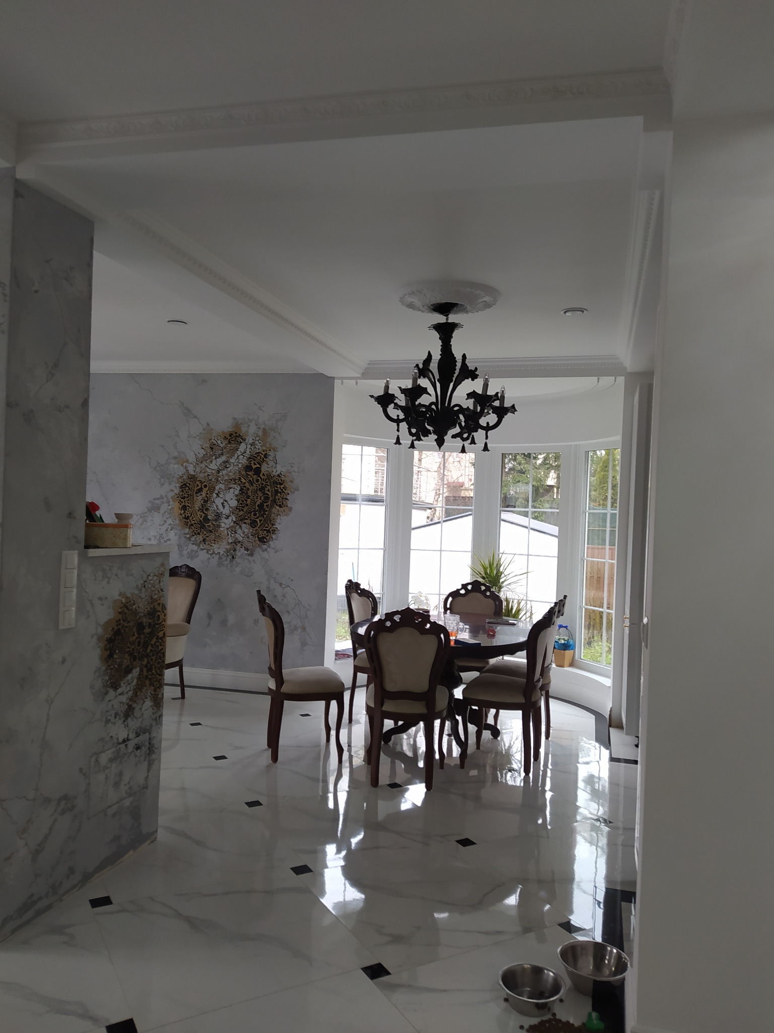 Eleganckie wnętrze jadalni z marmurową podłogą, dekoracyjną ścianą z motywem mandali, klasycznym żyrandolem i ciemnym stołem z krzesłami w stylu vintage, widoczne miski dla zwierząt.