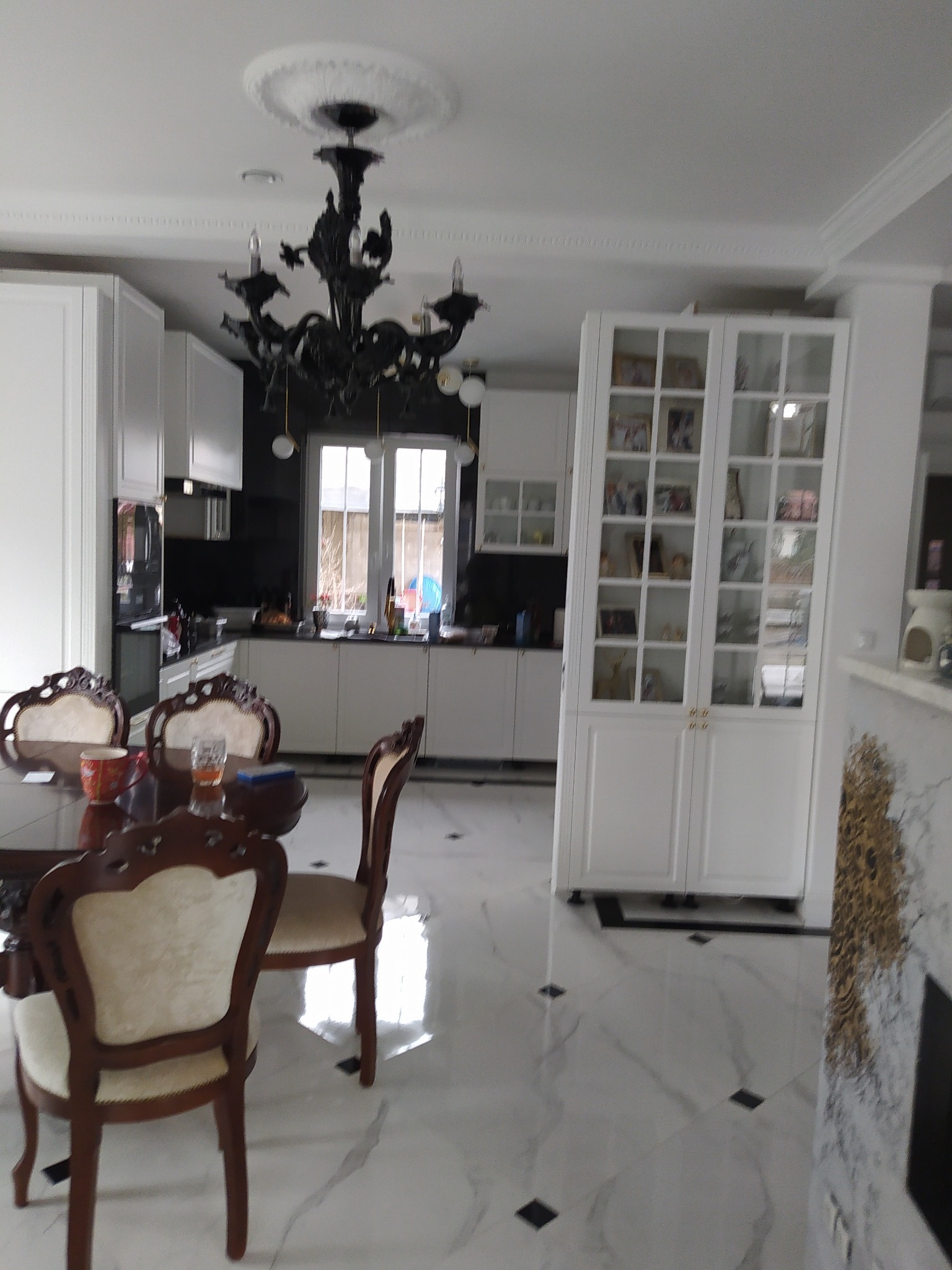 Jasne wnętrze jadalni z marmurową podłogą, białymi szafkami kuchennymi, czarnym żyrandolem i ciemną ścianą w tle.