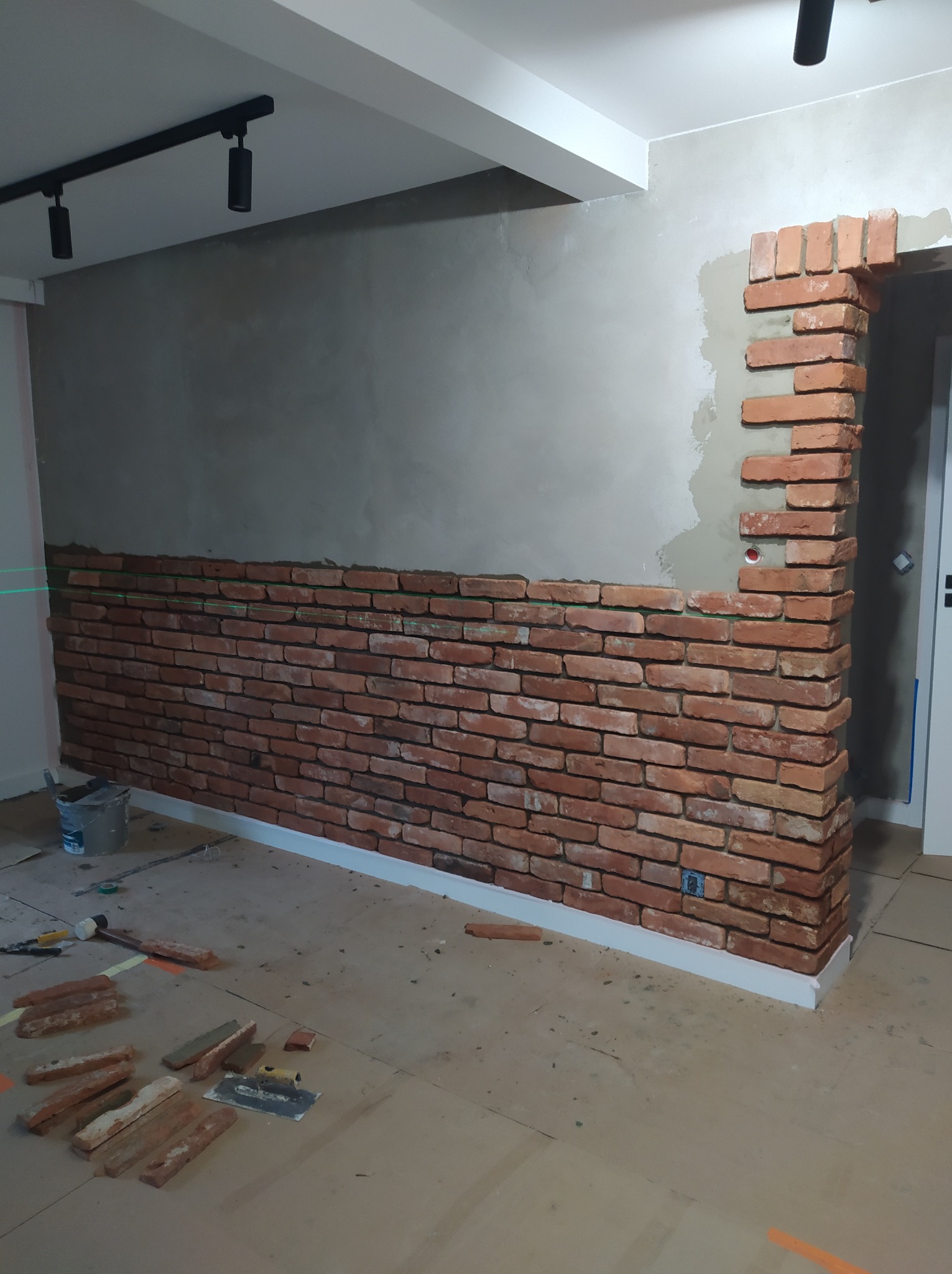 Montaż dekoracyjnej ściany z cegieł: widoczna ściana z częściowo ułożonymi cegłami, narzędzia pracy, poziomica laserowa, surowy beton na ścianie i czarne reflektory sufitowe.