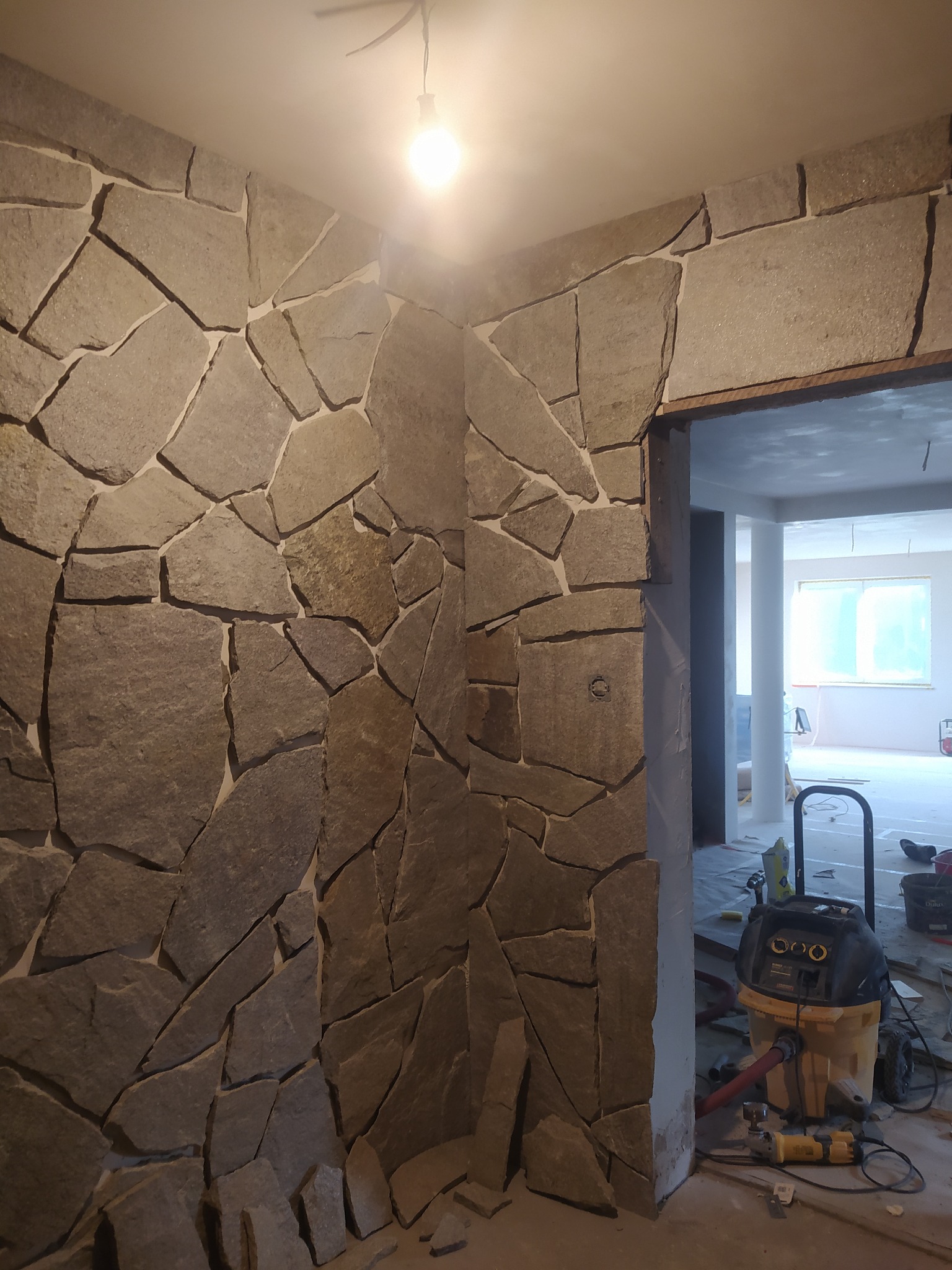 Ściana wykończona nieregularnymi płytami kamiennymi, widoczny odkurzacz przemysłowy i narzędzia przy wejściu do pomieszczenia w trakcie prac wykończeniowych.