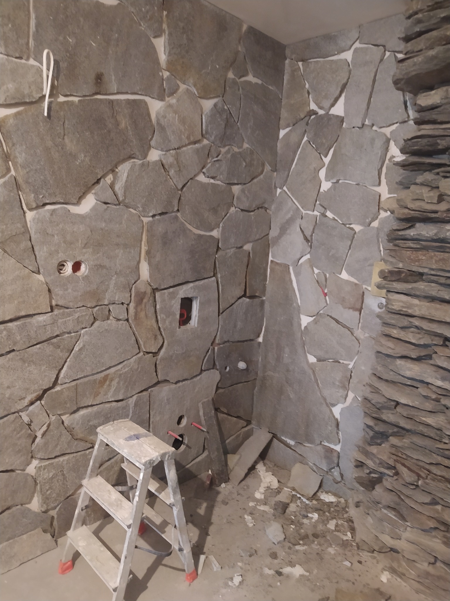 Ściana wykończona nieregularnymi kamiennymi płytami w odcieniach szarości, z widocznymi gniazdkami elektrycznymi i fragmentem ściany z wąskich, ułożonych warstwowo kamieni. Na podłodze stoi...