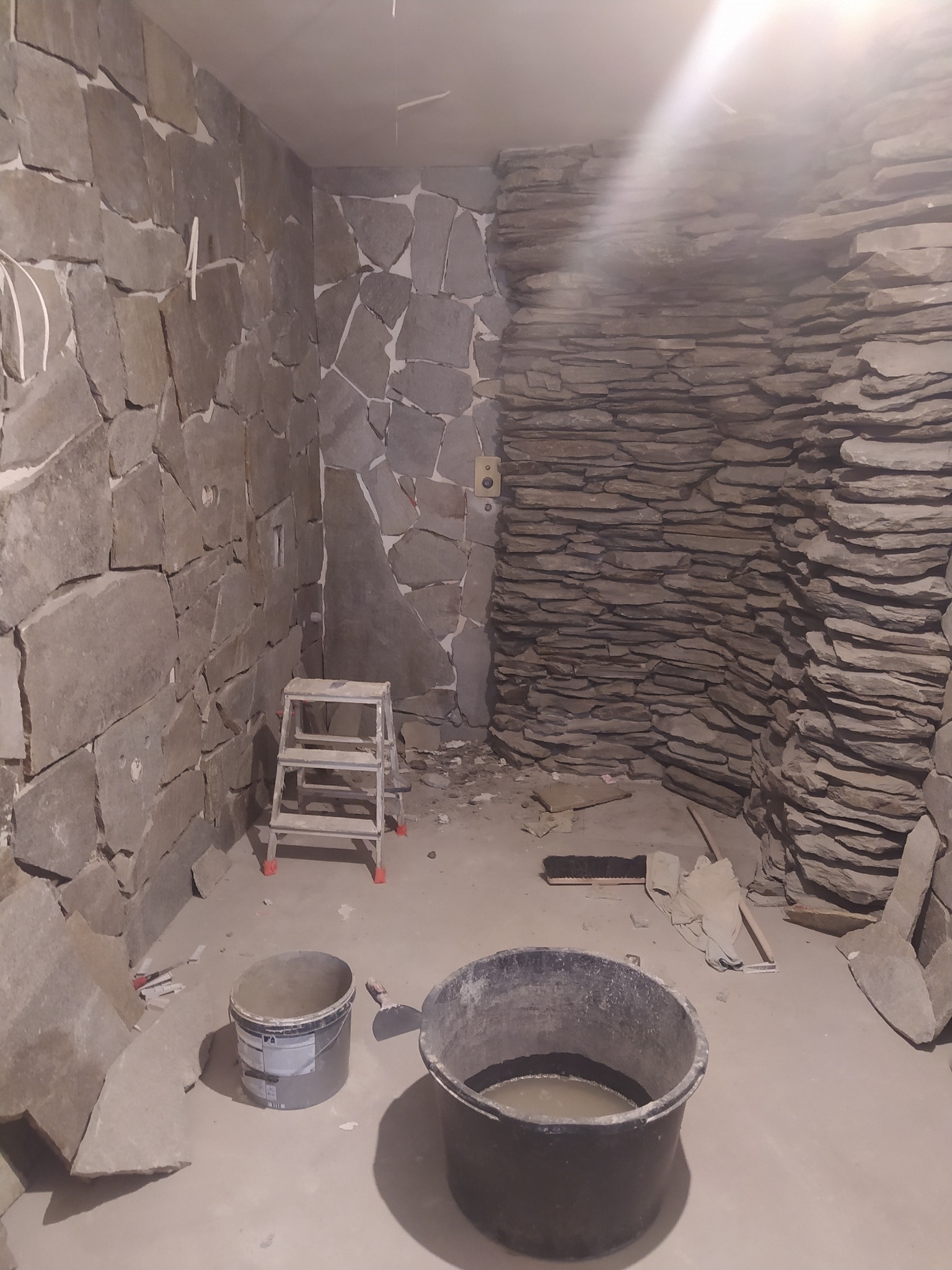 Wnętrze pomieszczenia w trakcie prac wykończeniowych, ściany pokryte kamieniem naturalnym w dwóch różnych stylach, widoczne wiadro z zaprawą, drabina i narzędzia.
