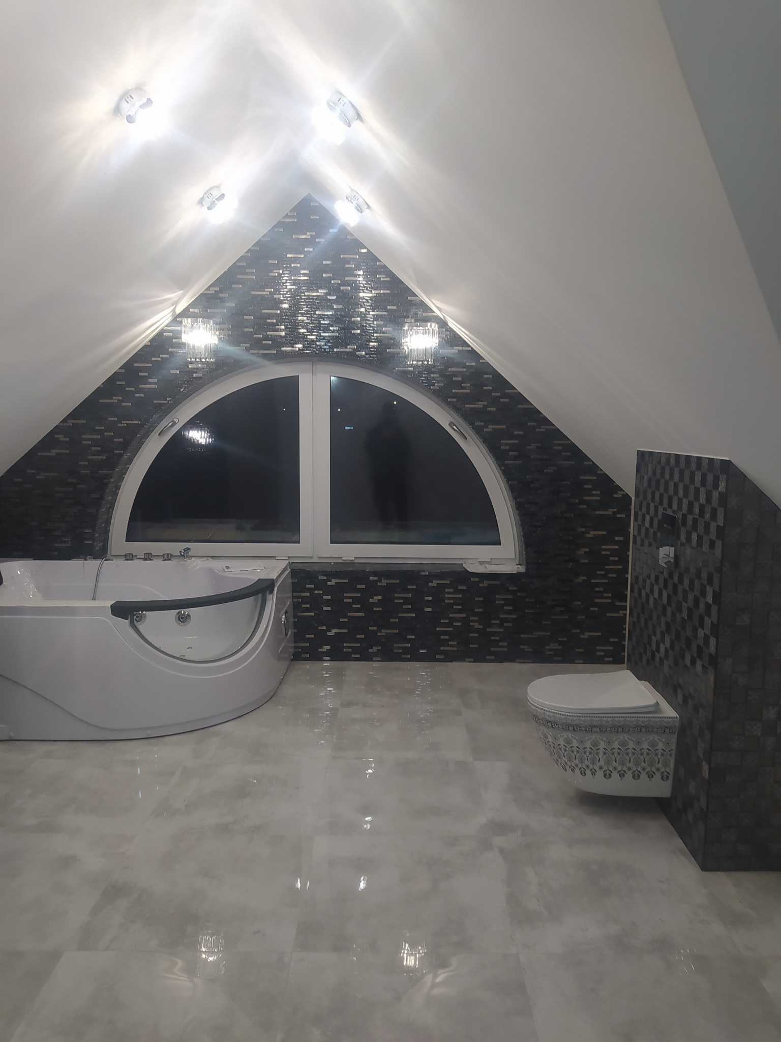 Wykończona łazienka na poddaszu ze spadzistym sufitem, mozaiką na ścianach, owalnym oknem, białą wanną z hydromasażem i podwieszaną toaletą z dekoracyjnym wzorem.