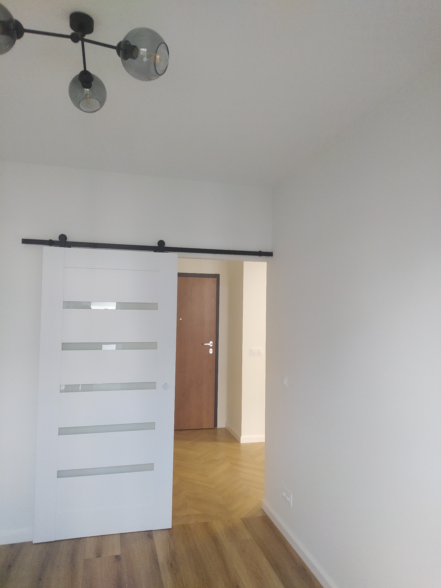 Białe przesuwne drzwi z poziomymi szybami zamontowane na czarnej szynie, widok na parkiet w jodełkę i drzwi wejściowe w tle, nowoczesne oświetlenie sufitowe.