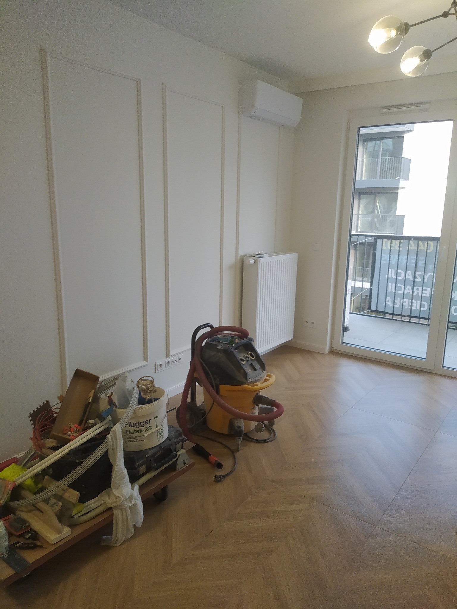 Wnętrze w trakcie prac wykończeniowych: białe ściany z dekoracyjnymi listwami, parkiet w jodełkę, grzejnik, klimatyzacja oraz wózek z narzędziami i odkurzacz przemysłowy.