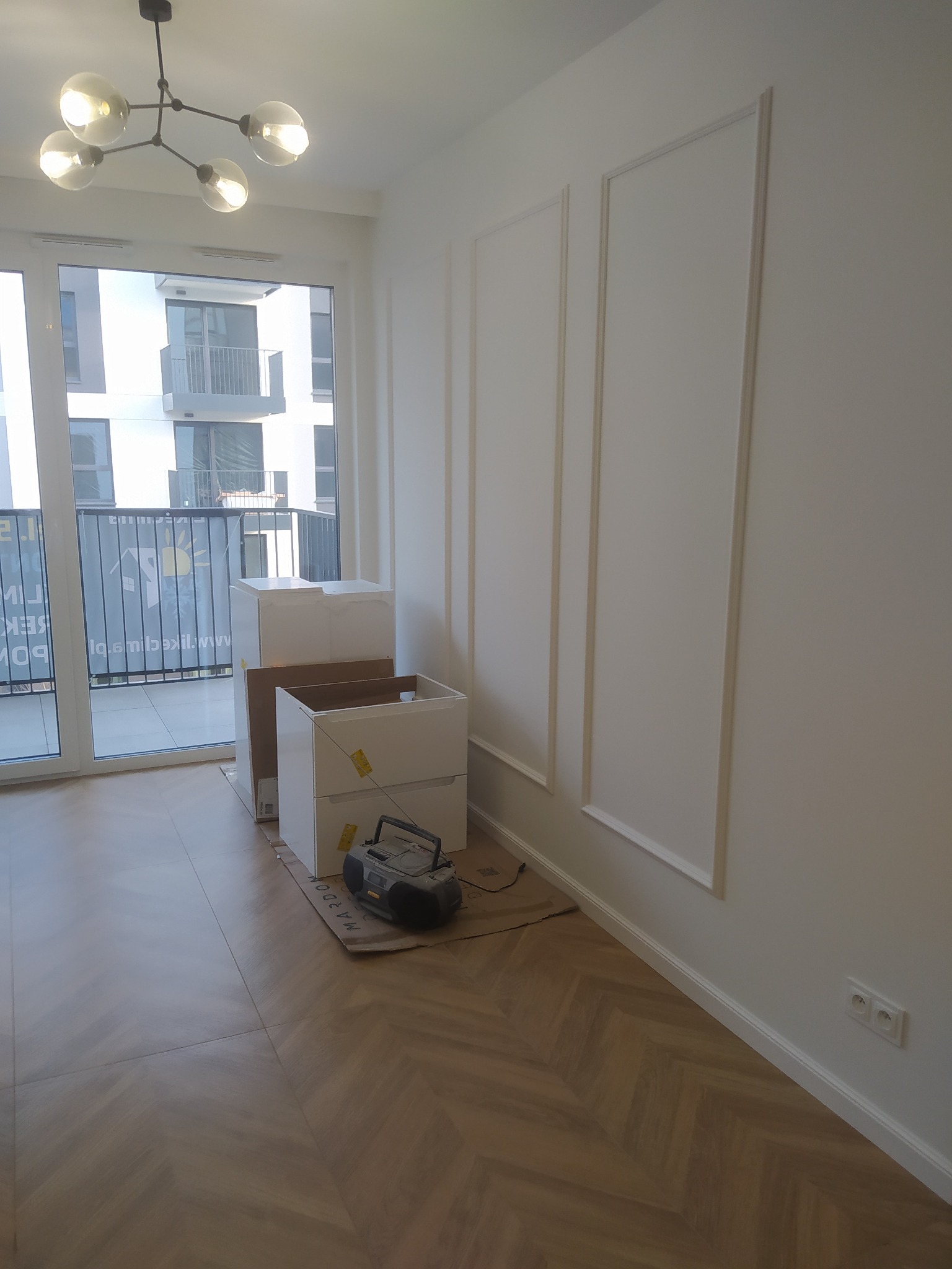 Wnętrze mieszkania w trakcie prac wykończeniowych: białe ściany z dekoracyjnymi listwami, podłoga w jodełkę, niezmontowane białe szafki w kartonach i radio na podłodze, widok z okna na budynek...