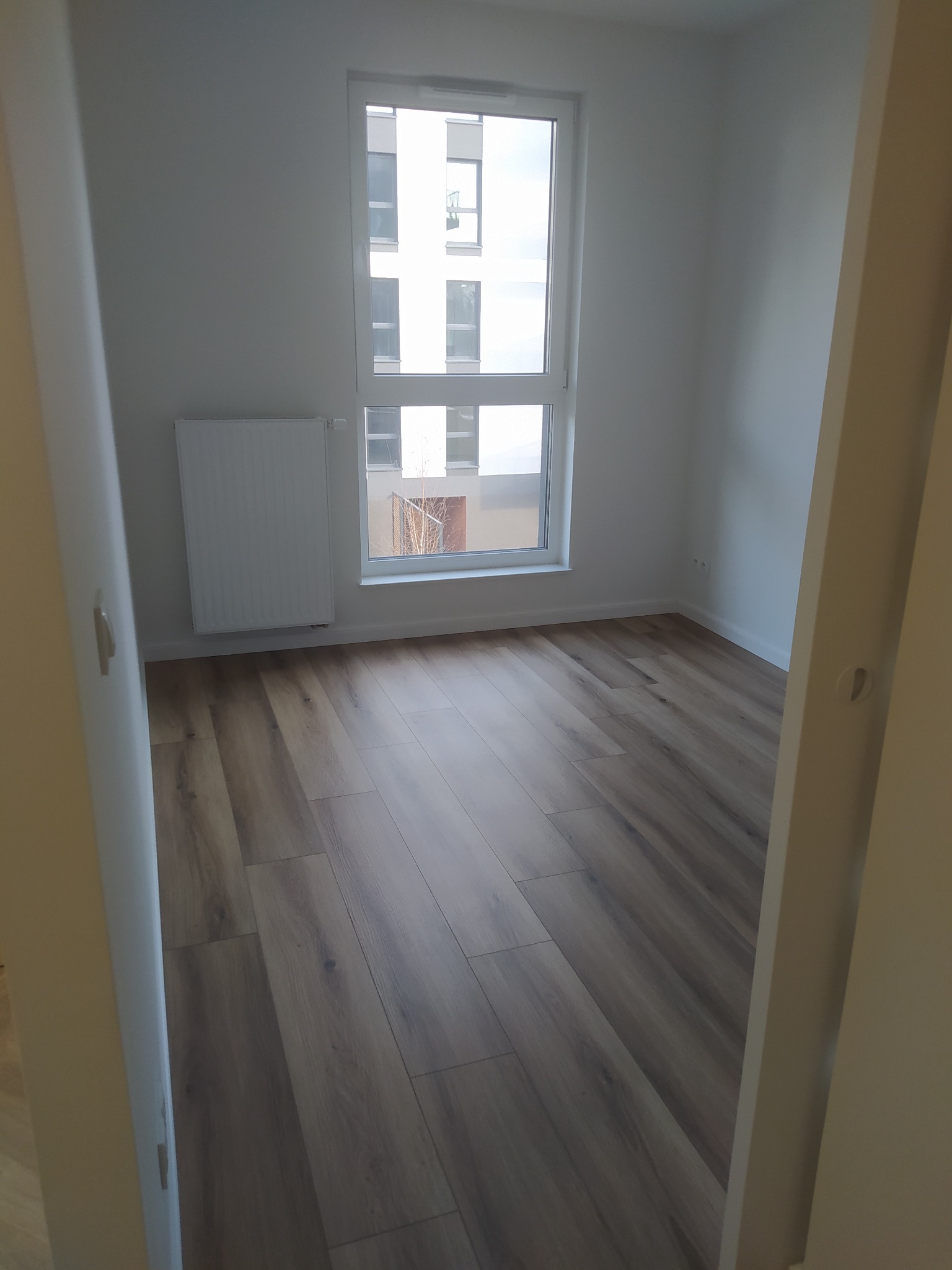 Pusty pokój z panelami podłogowymi w odcieniach brązu, białymi ścianami i grzejnikiem pod oknem, widok zza framugi drzwi.