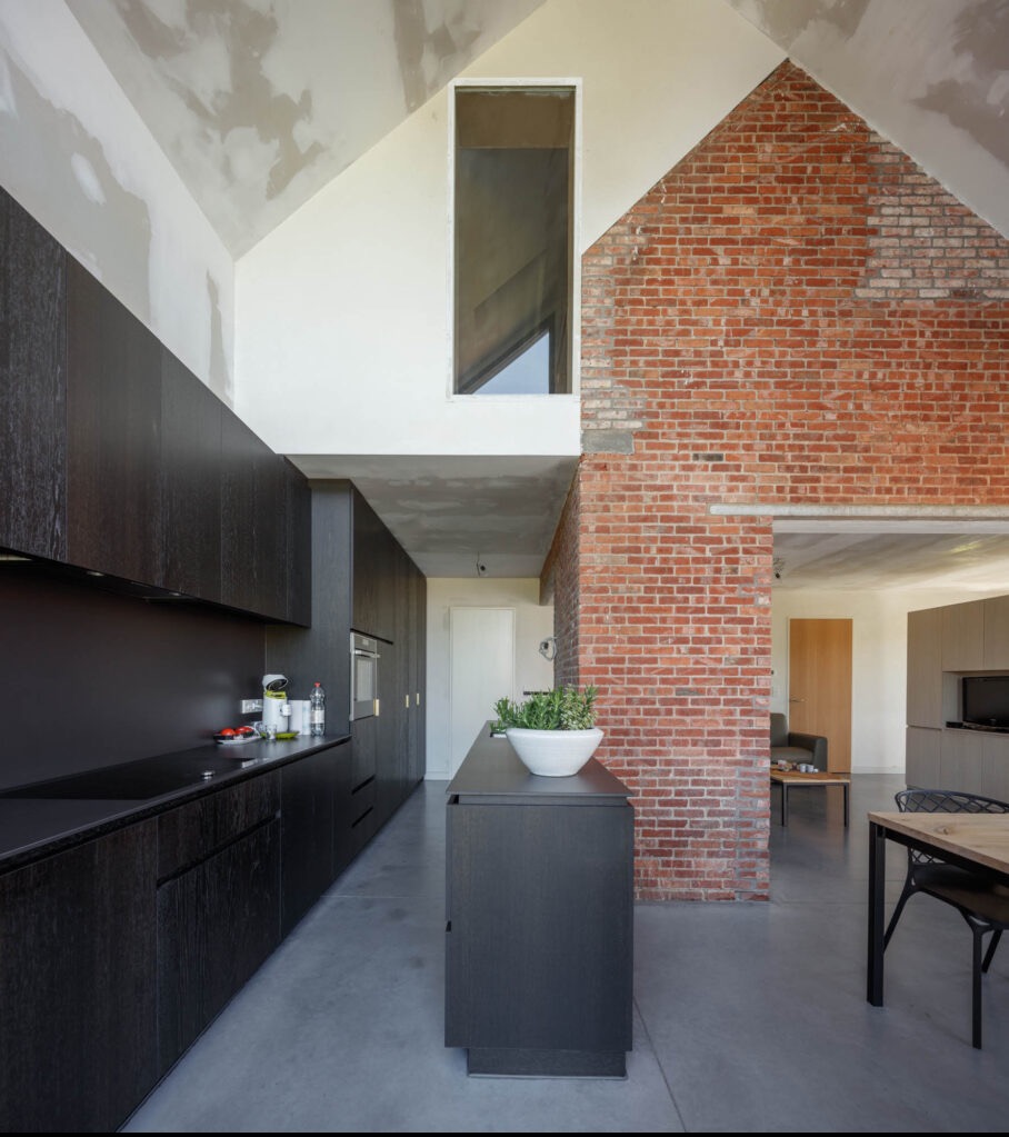 Nowoczesna kuchnia z ciemnymi, drewnianymi szafkami, surową ceglaną ścianą i wysokim oknem w kształcie pionowego prostokąta, sufit częściowo wykończony gładzią, widok na dalszą część domu.