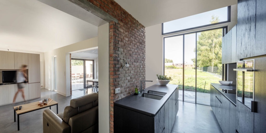 Nowoczesna kuchnia z wyspą i ceglaną ścianą, duże okno z widokiem na zieleń, w tle salon z osobą w ruchu, minimalistyczny design.