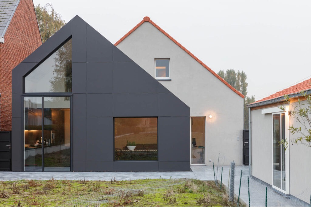 Nowoczesny dom z dobudowaną, ciemnoszarą częścią o geometrycznym kształcie, z dużymi oknami, kontrastujący z jasnym, tradycyjnym budynkiem z czerwonym dachem.