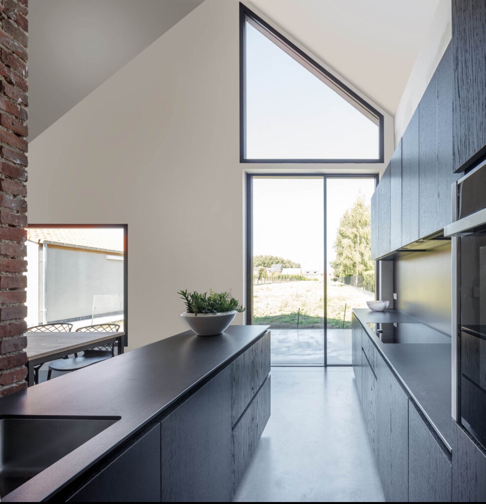 Nowoczesna kuchnia w domu z wysokim oknem trójkątnym, czarne matowe szafki z widoczną strukturą drewna, ceglany filar, widok na zewnątrz przez przesuwne drzwi.