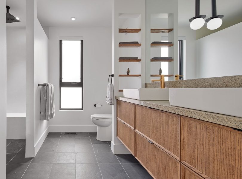 Nowoczesna łazienka z dwoma białymi umywalkami na drewnianej szafce z kamiennym blatem, półki z drewnianymi deskami i szarymi ręcznikami, czarne płytki podłogowe i okno z czarną ramą.