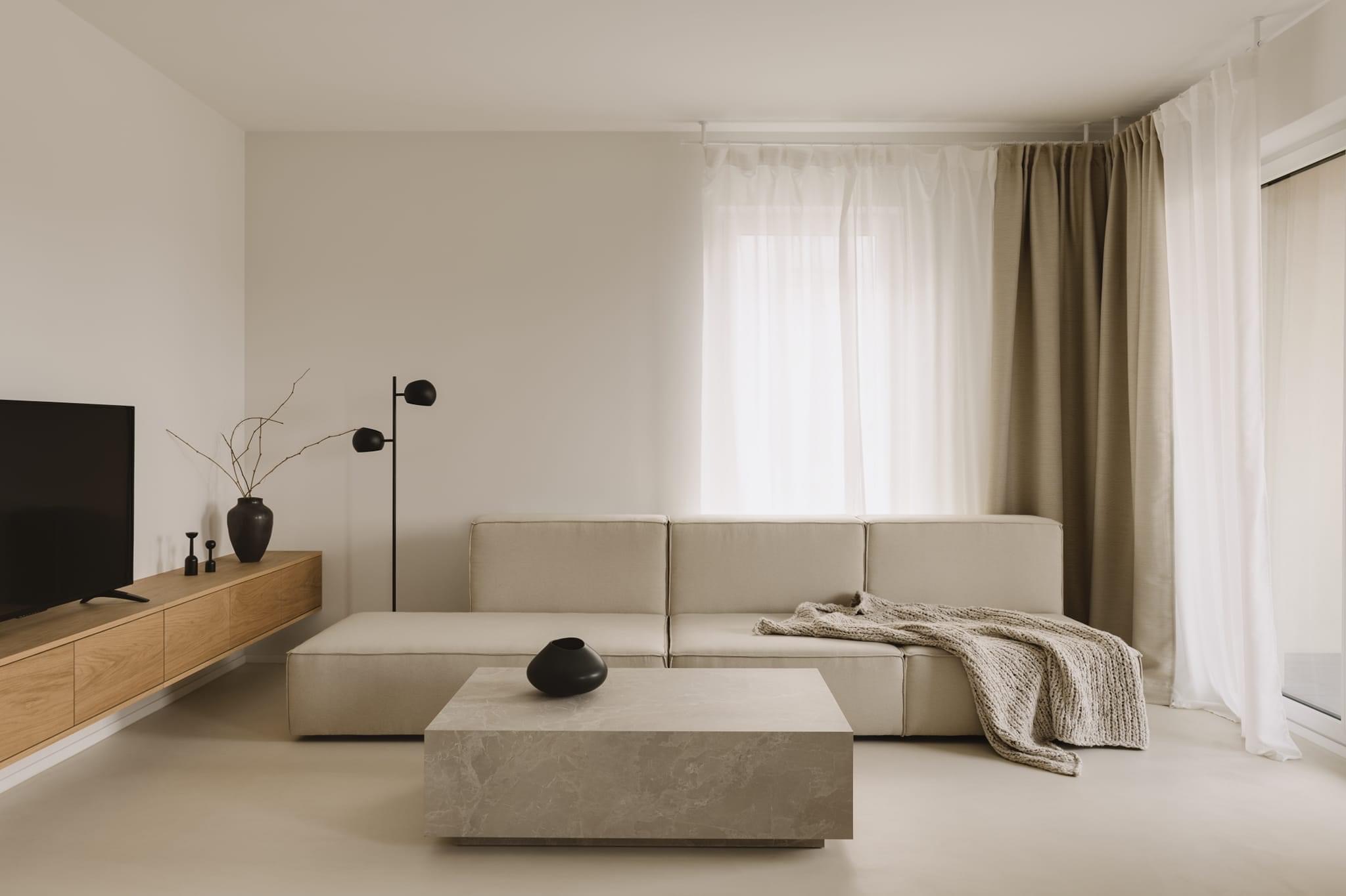 Minimalistyczny salon z beżową sofą modułową, marmurkowym stolikiem kawowym, drewnianą szafką RTV i czarnymi dodatkami, oświetlony naturalnym światłem wpadającym przez okno z białymi i beżowymi...