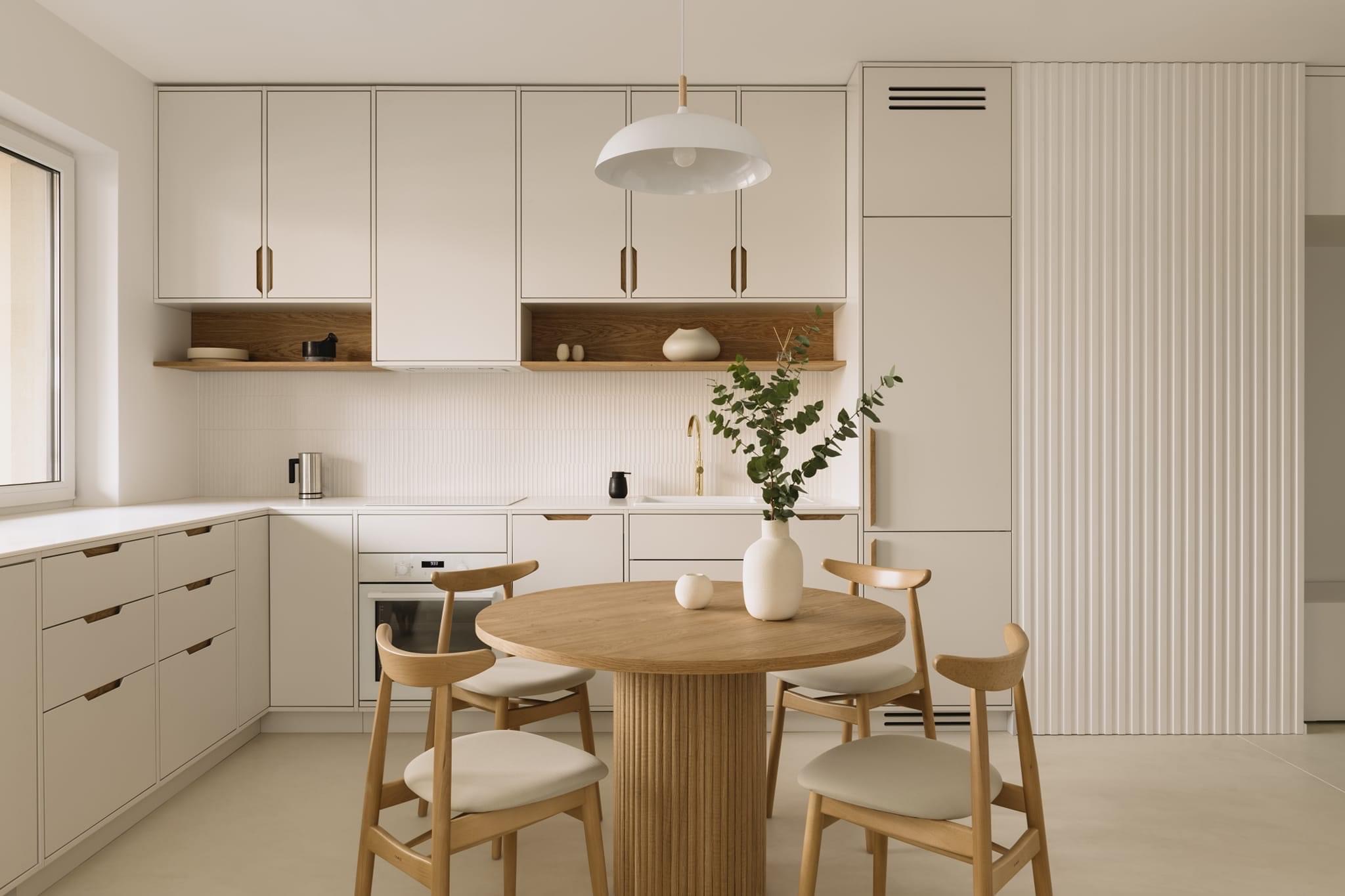 Jasna kuchnia w minimalistycznym stylu z okrągłym drewnianym stołem i krzesłami, białe szafki z drewnianymi uchwytami, białe płytki na ścianie i dekoracyjne panele ścienne.
