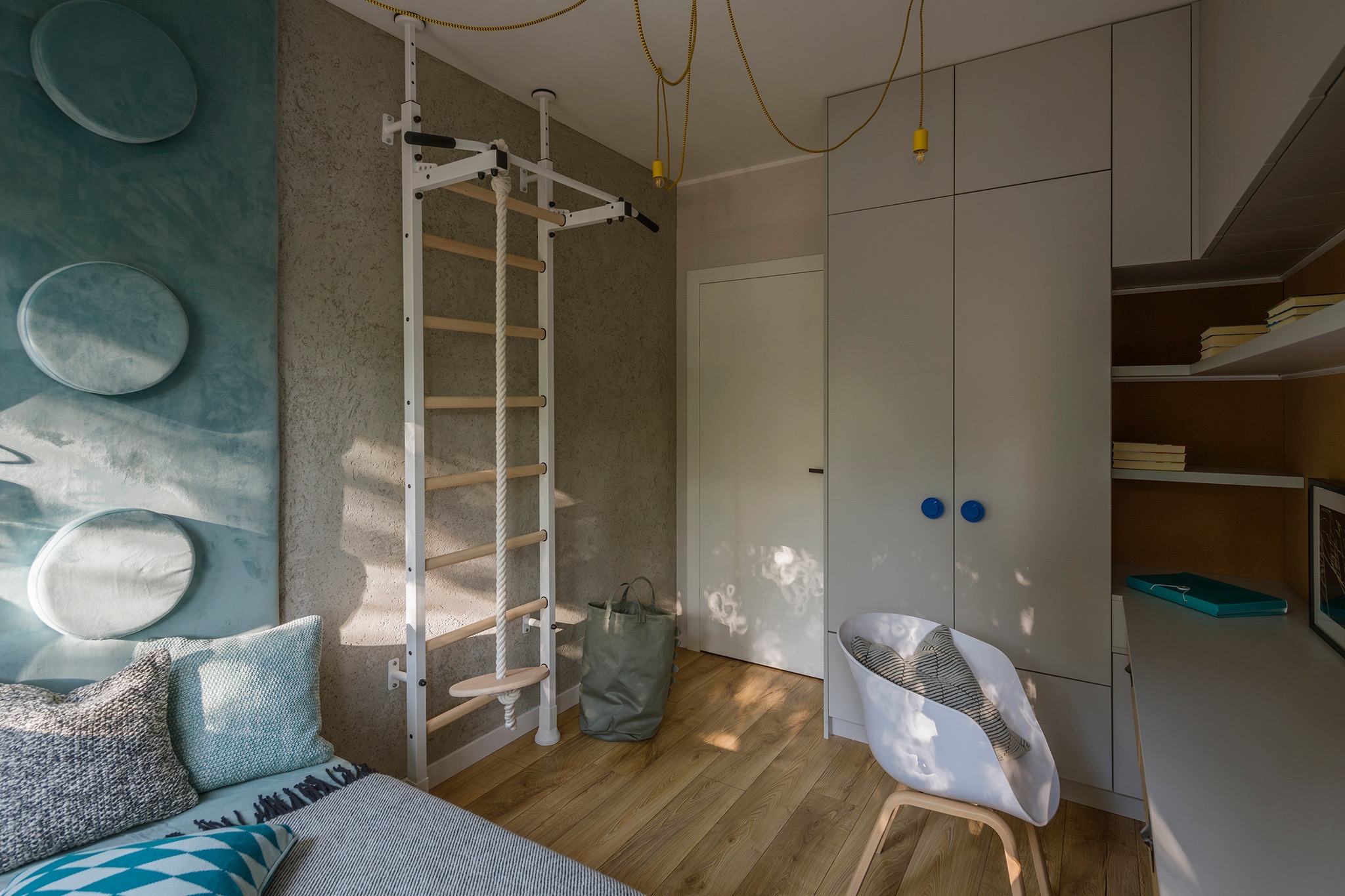 Pokój dziecięcy z drewnianą podłogą, drabinką gimnastyczną z liną, łóżkiem z okrągłymi poduszkami na ścianie, szarą szafą z niebieskimi uchwytami i białym krzesłem z poduszką w kształcie ryby.