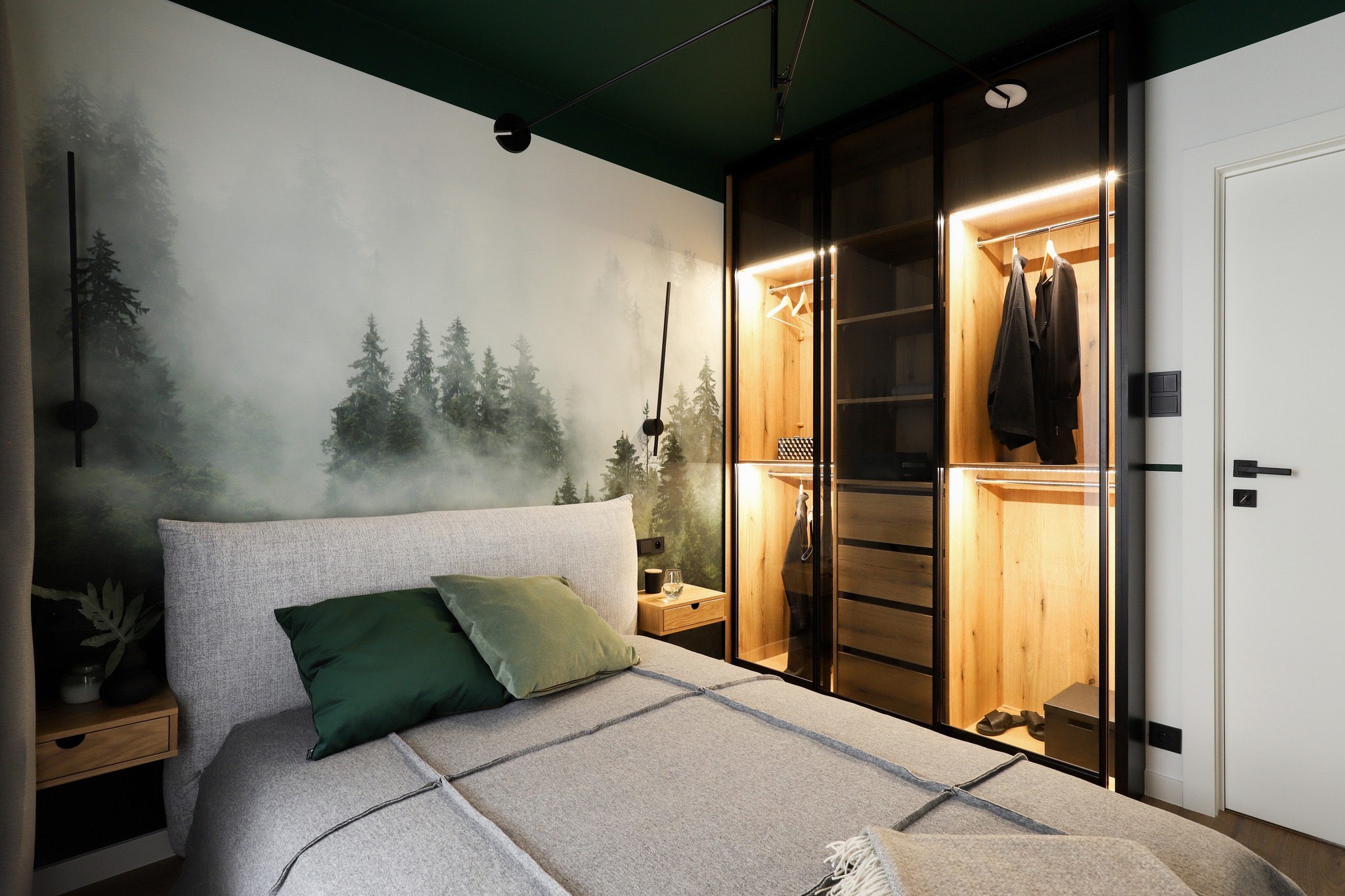 Sypialnia z łóżkiem z szarą narzutą i zielonymi poduszkami, tapetą z motywem leśnym za łóżkiem, szafkami nocnymi po bokach i podświetlaną szafą przesuwną z czarnymi ramami.
