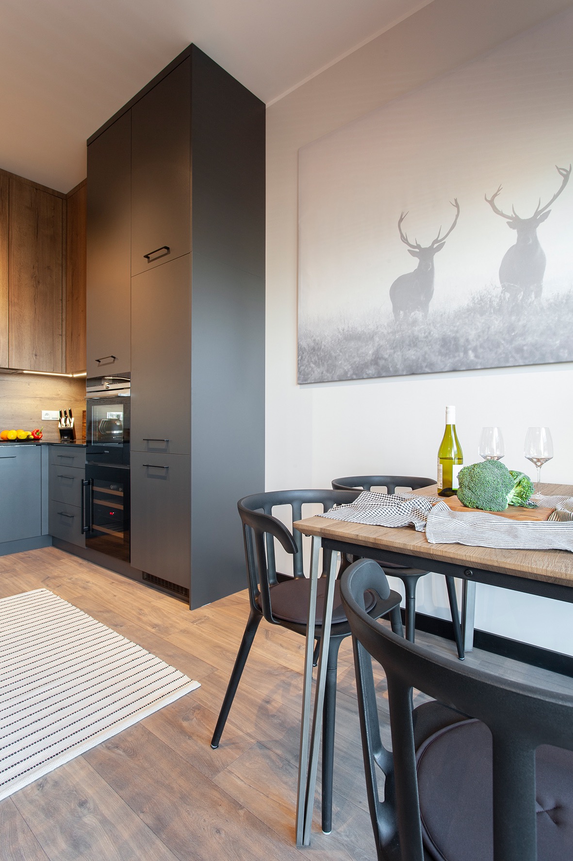 Nowoczesna kuchnia z drewnianymi i grafitowymi szafkami, drewnianym stołem z czarnymi krzesłami, butelką wina i brokułami, oraz obrazem z motywem jeleni na ścianie.