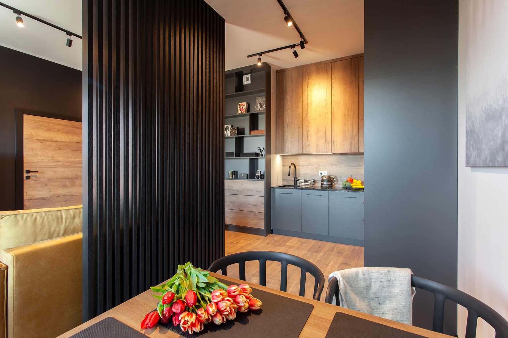 Nowoczesne wnętrze: kuchnia z szarymi szafkami i drewnianymi akcentami, oddzielona od salonu czarną, pionową przegrodą. Na pierwszym planie stół z bukietem czerwonych tulipanów.