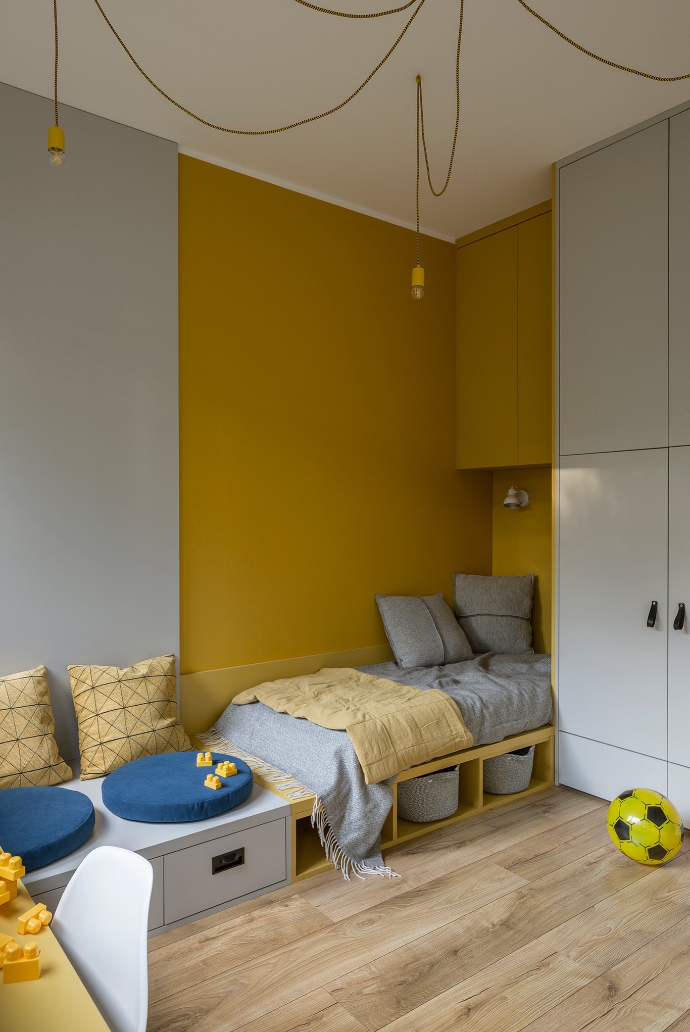 Pokój dziecięcy z zabudowanym łóżkiem, szarą szafą wnękową, żółtą ścianą i wiszącymi lampami na złotych kablach, uzupełniony niebieskimi pufami i żółtą piłką.