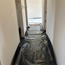 INSTAL-PARTNER Technika Grzewcza i Sanitarna Radosław Hązła - Korytarz w trakcie przygotowania do wylewki pod ogrzewanie podłogowe, zabezpieczony czarną folią, widoczne rolki folii i białe płyty izolacyjne w oddali.