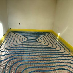 INSTAL-PARTNER Technika Grzewcza i Sanitarna Radosław Hązła - Niebieskie rury ogrzewania podłogowego ułożone spiralnie na folii z czerwoną siatką, żółta taśma brzegowa przy ścianach, widoczne kable elektryczne.