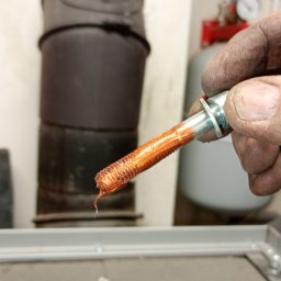 INSTAL-PARTNER Technika Grzewcza i Sanitarna Radosław Hązła - Brudna dłoń hydraulika trzyma śrubę pokrytą uszczelniaczem do gwintów, w tle piec CO, rury instalacji i manometr.