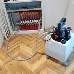 INSTAL-PARTNER Technika Grzewcza i Sanitarna Radosław Hązła - Rozdzielacz ogrzewania podłogowego w szafce, podłączony do pompy ciśnieniowej do napełniania i odpowietrzania instalacji, widoczne rury i zawory.