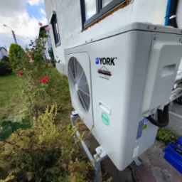 Biała pompa ciepła YORK zamontowana na zewnątrz budynku, widoczna z boku, z narzędziami w niebieskiej skrzynce obok. W tle ogród z kwitnącymi różami.