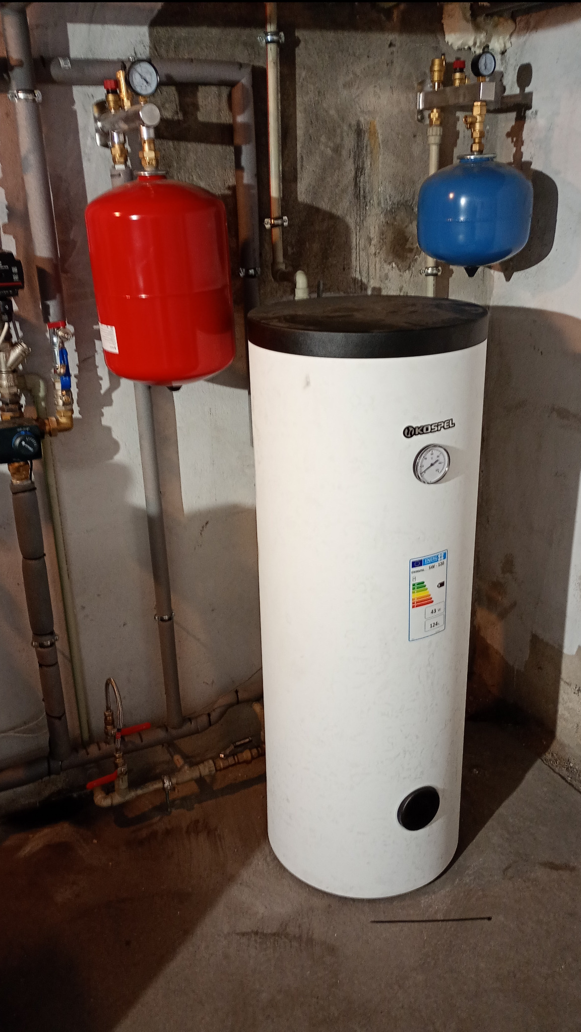Bojler Kospel w piwnicy z podłączonymi instalacjami, widoczny manometr i etykieta energetyczna.