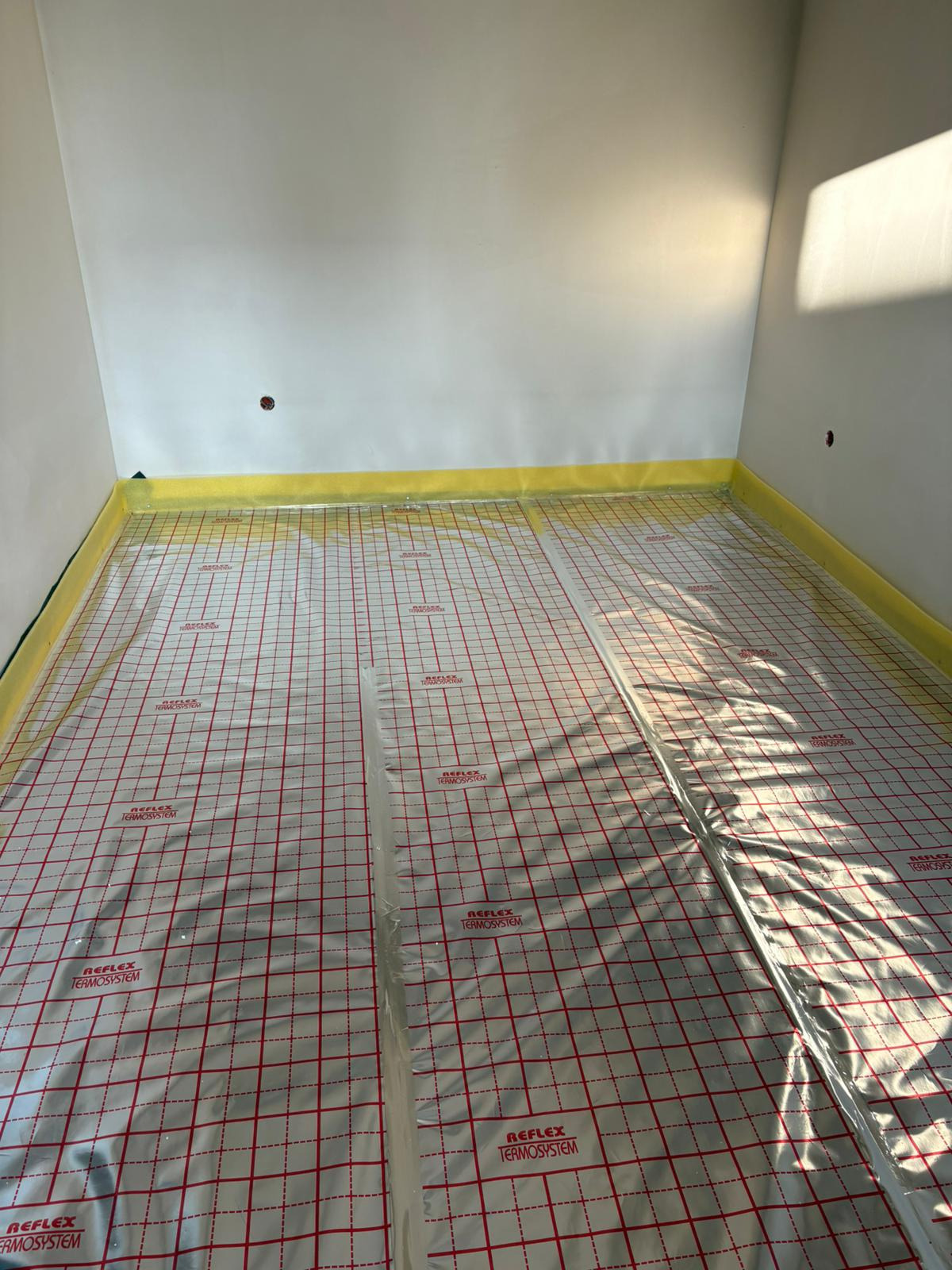 Przygotowana powierzchnia pod wylewkę ogrzewania podłogowego z folią refleksyjną w pomieszczeniu z żółtą taśmą zabezpieczającą przy ścianach.
