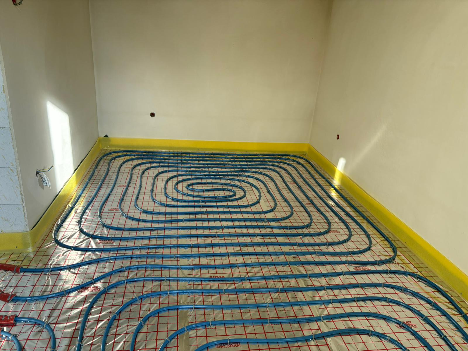 Niebieskie rury ogrzewania podłogowego ułożone spiralnie na folii z czerwoną siatką, żółta taśma brzegowa przy ścianach, widoczne kable elektryczne.