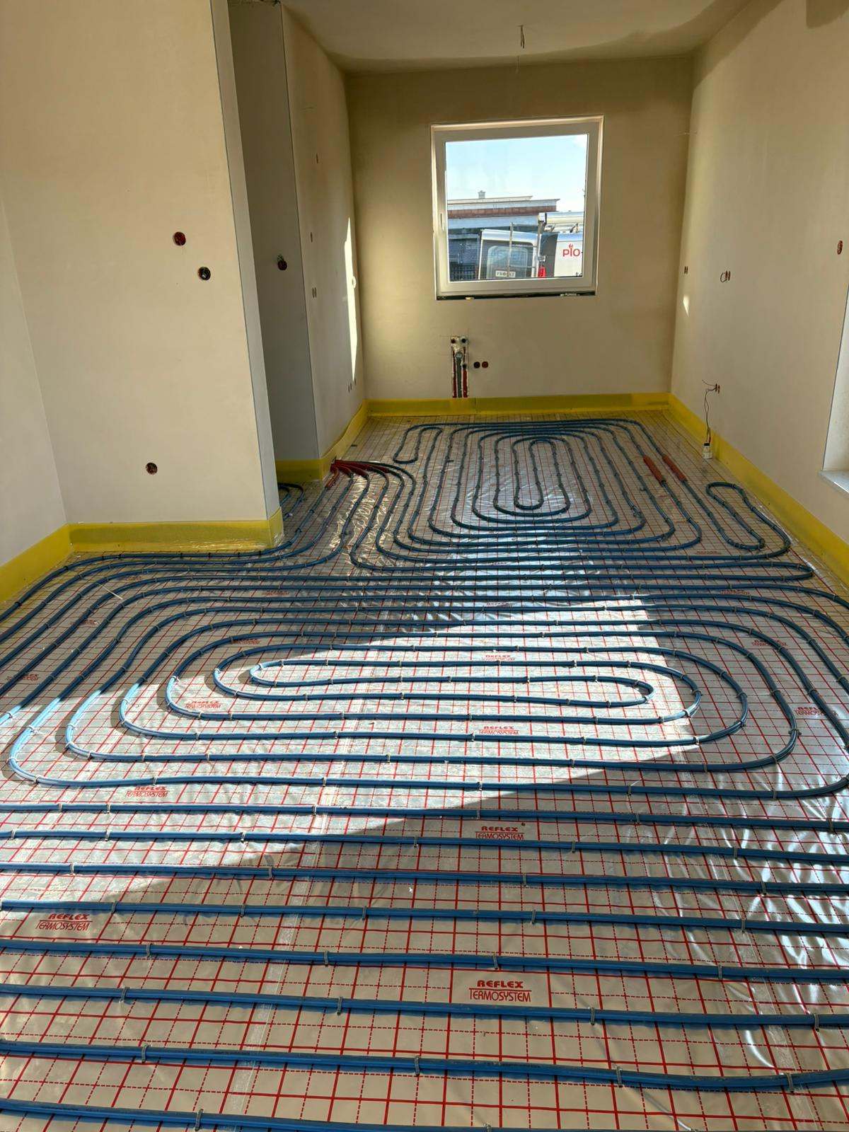 Niebieskie rury ogrzewania podłogowego ułożone na czerwonej siatce w surowym pomieszczeniu z oknem i przygotowanymi instalacjami na ścianach.