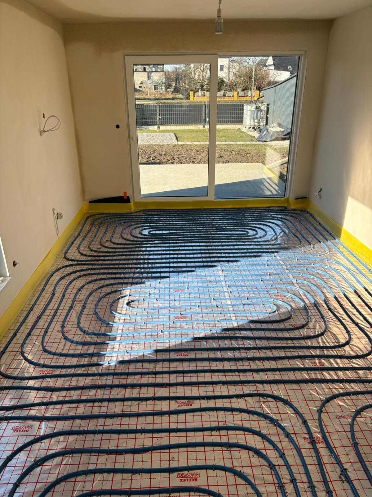 Rozłożone rury ogrzewania podłogowego na folii z siatką w surowym stanie, widoczne okno z widokiem na ogród.