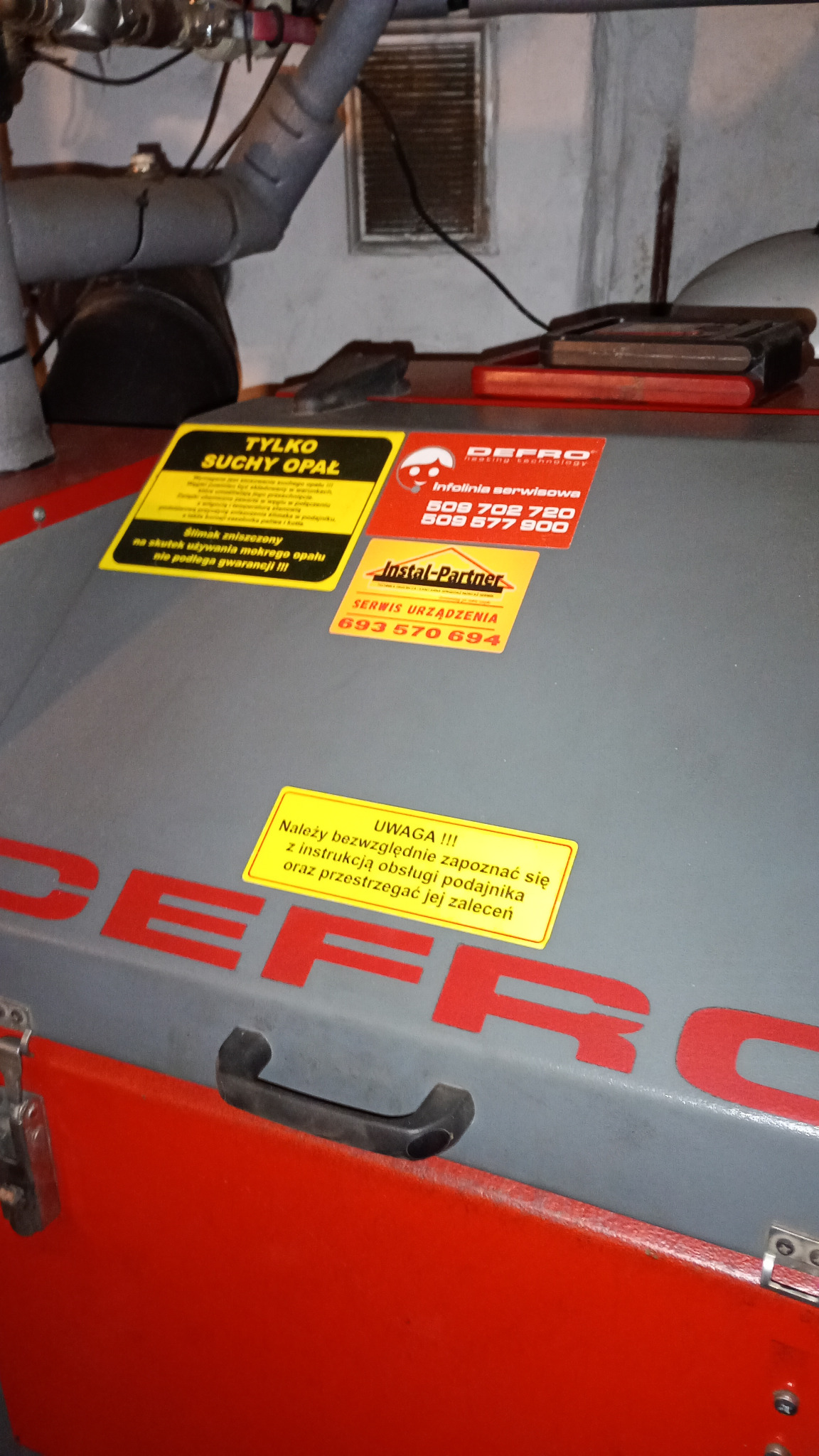 Piec centralnego ogrzewania marki DEFRO z naklejonymi informacjami o serwisie i ostrzeżeniami dotyczącymi używania suchego opału.