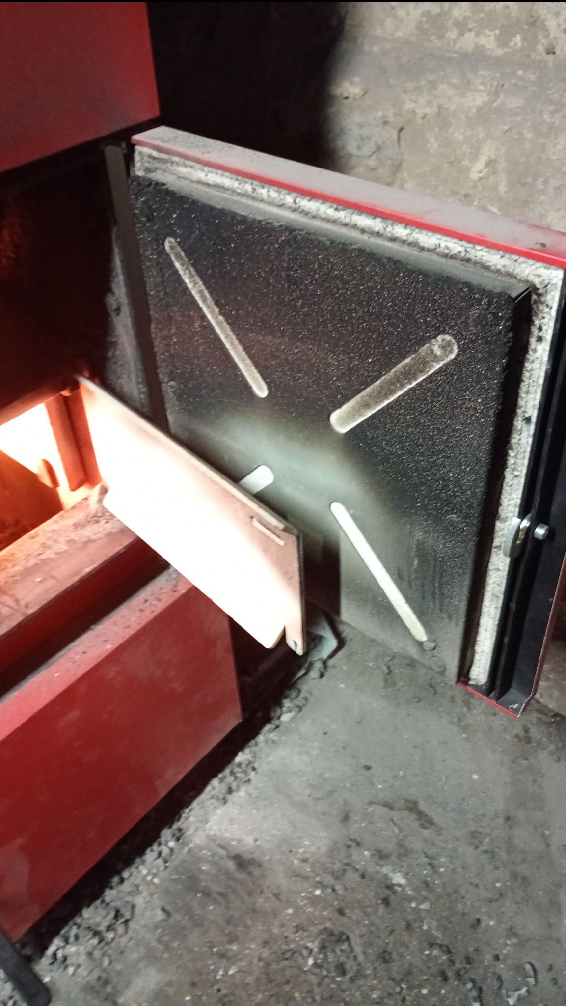 Otwarta, brudna klapa pieca CO z widocznym paleniskiem i żarem wewnątrz, na betonowej podłodze.