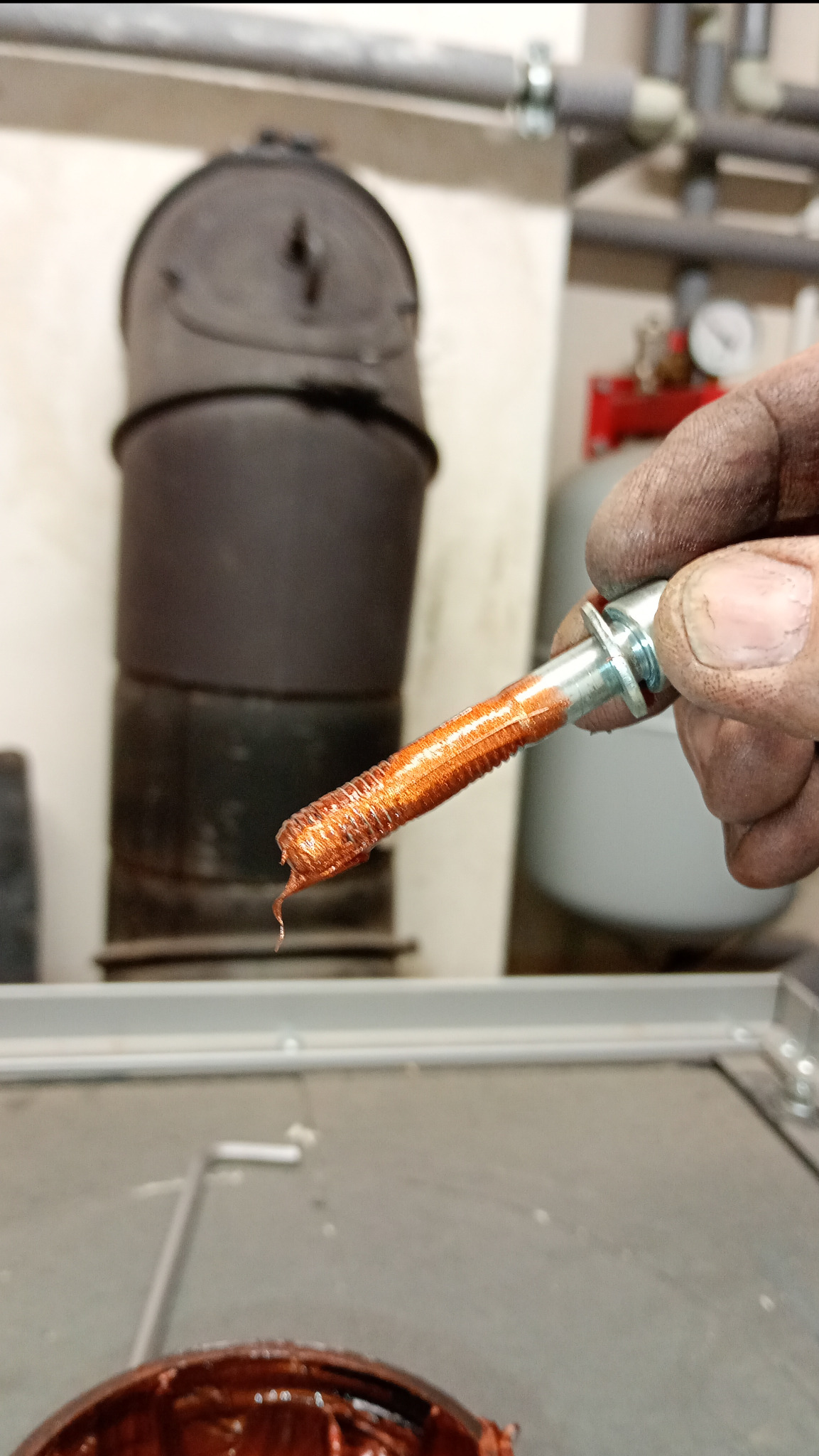 Brudna dłoń hydraulika trzyma śrubę pokrytą uszczelniaczem do gwintów, w tle piec CO, rury instalacji i manometr.