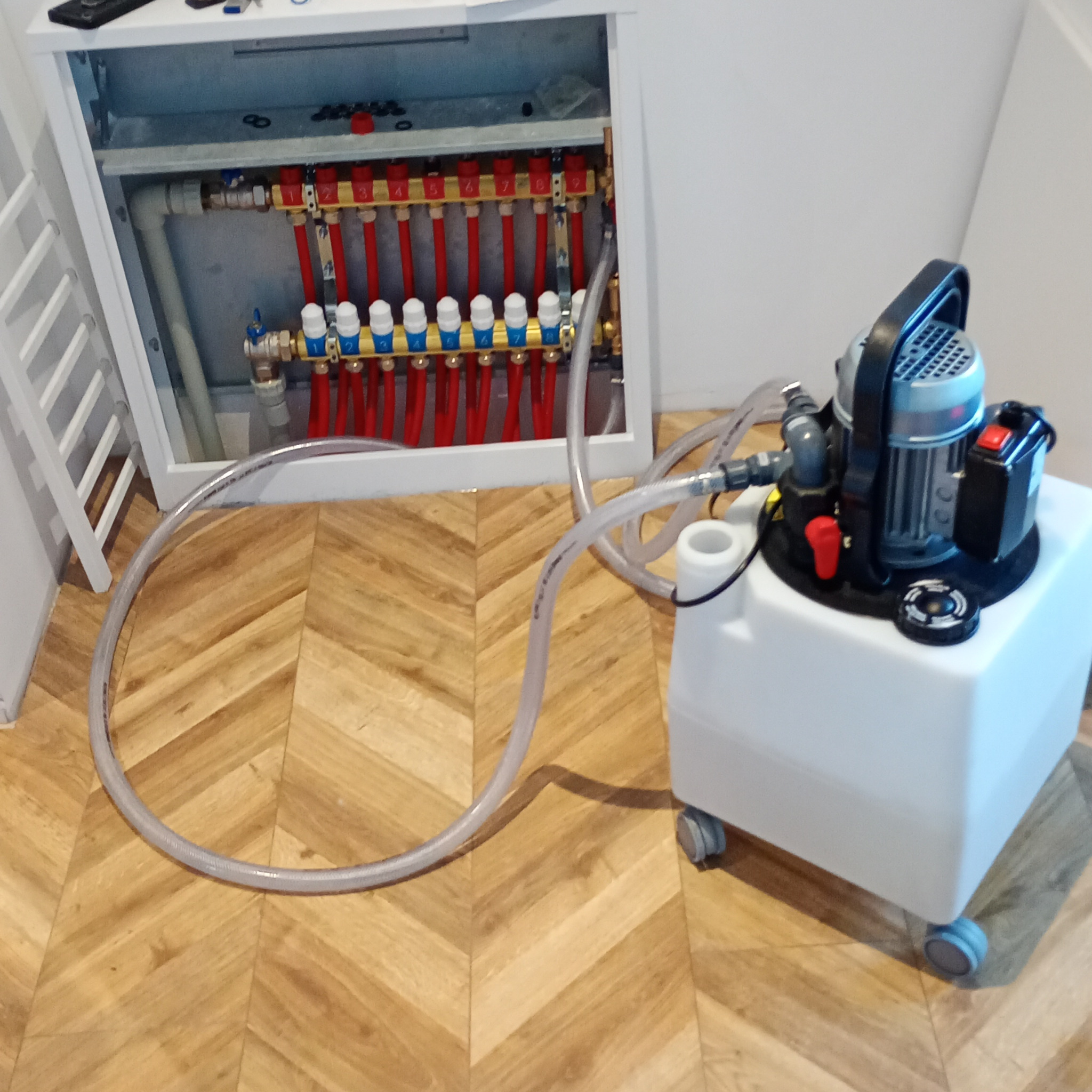 Rozdzielacz ogrzewania podłogowego w szafce, podłączony do pompy ciśnieniowej do napełniania i odpowietrzania instalacji, widoczne rury i zawory.