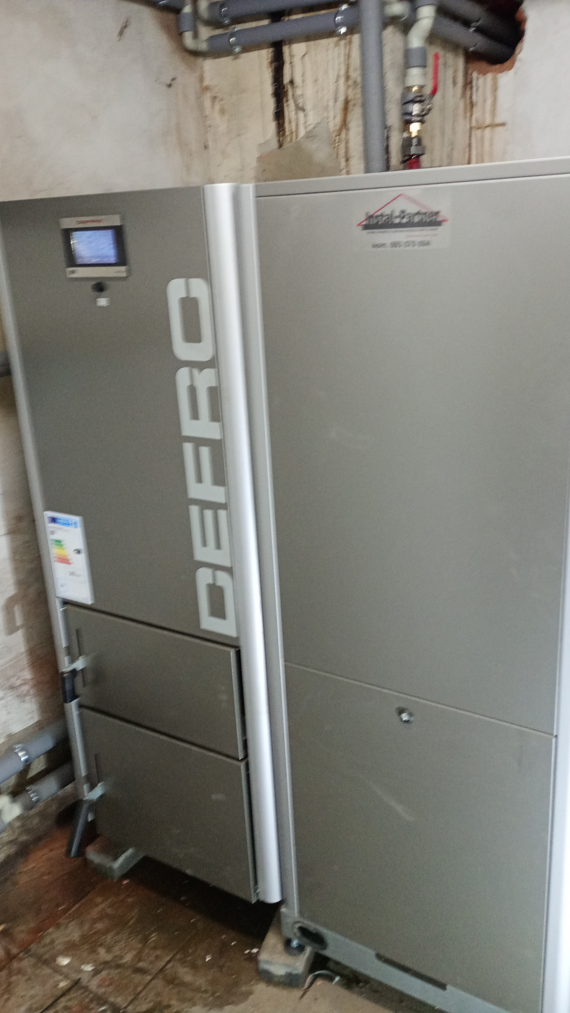 Szary piec CO marki Defro z wyświetlaczem i etykietą energetyczną, widoczne rury instalacji grzewczej na tle ściany z zaciekami.