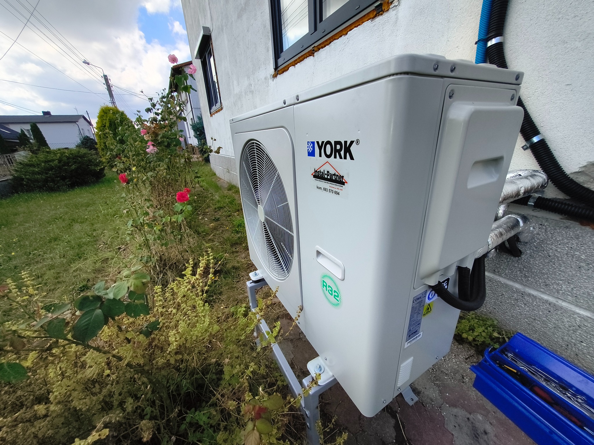 Biała pompa ciepła YORK zamontowana na zewnątrz budynku, widoczna z boku, z narzędziami w niebieskiej skrzynce obok. W tle ogród z kwitnącymi różami.