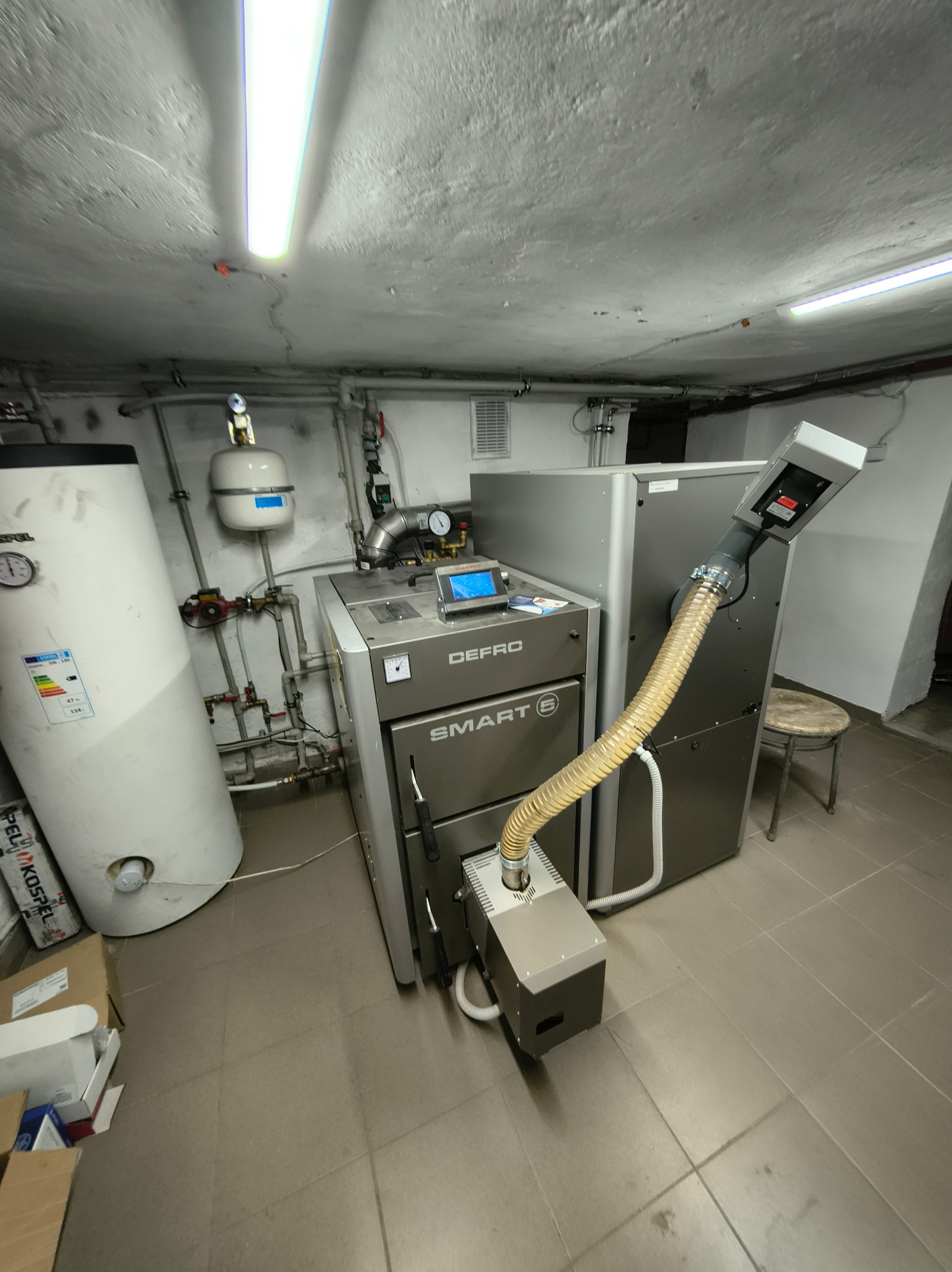 Szary piec Defro Smart 5 z podajnikiem w kotłowni, obok biały zbiornik na wodę i szare elementy instalacji. Widoczne rury, zawory i manometry.