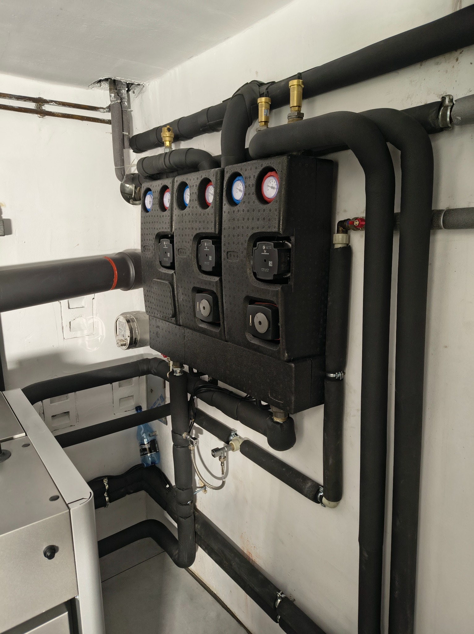 Czarna, zaizolowana instalacja grzewcza z manometrami i zaworami, zamontowana na białej ścianie w kotłowni. Widoczne rury i elementy pomiarowe.