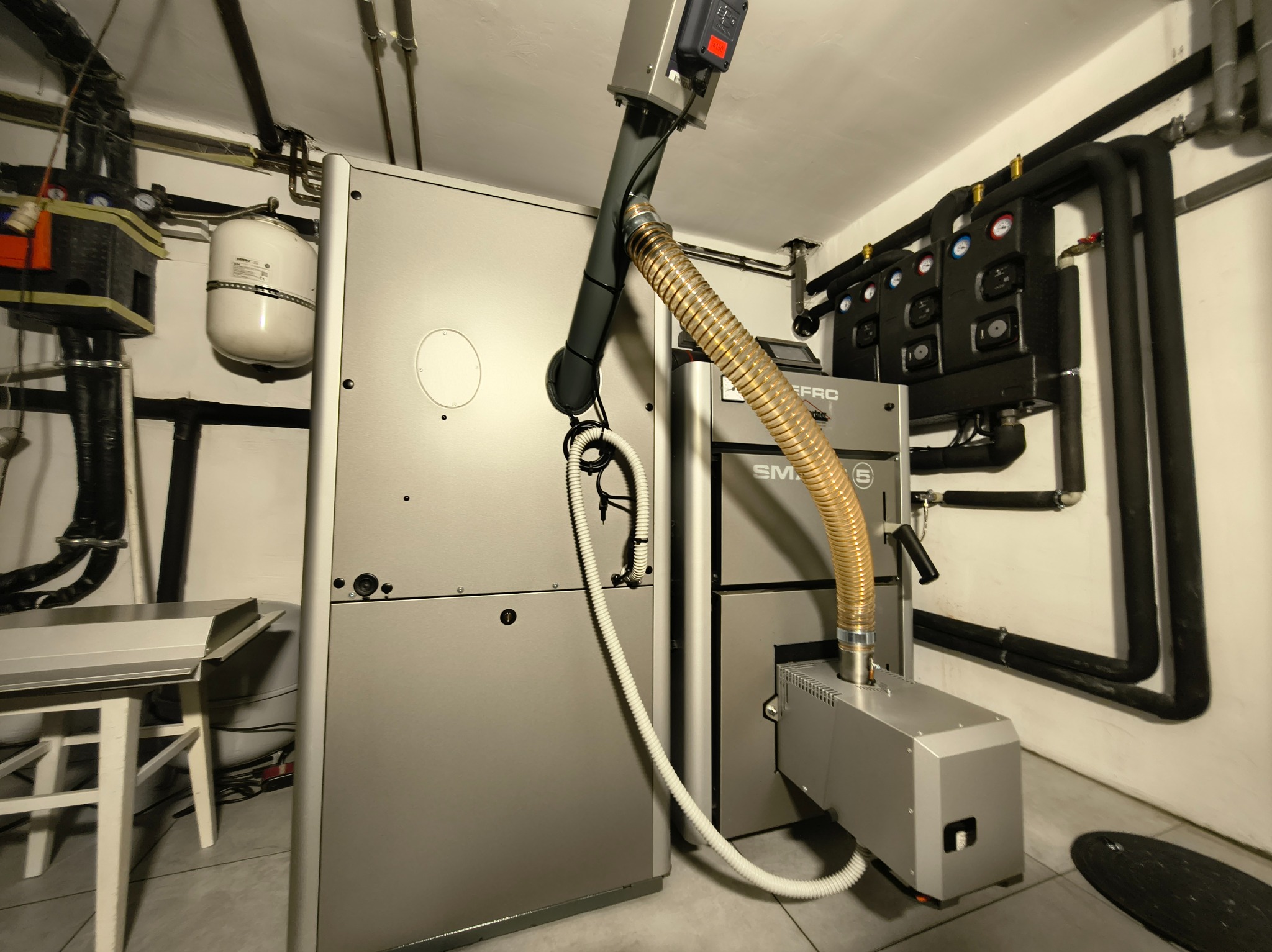 Nowoczesny piec CO na ekogroszek w kotłowni, widoczne rury instalacji grzewczej i zbiornik wyrównawczy. Oświetlenie sufitowe. Szare płytki na podłodze.